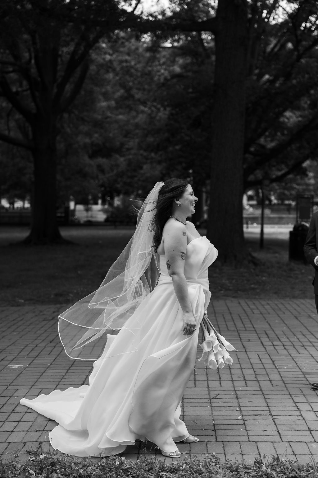 Bride in strapless white gown and flowing veil walks through a park holding tulips on her wedding day.