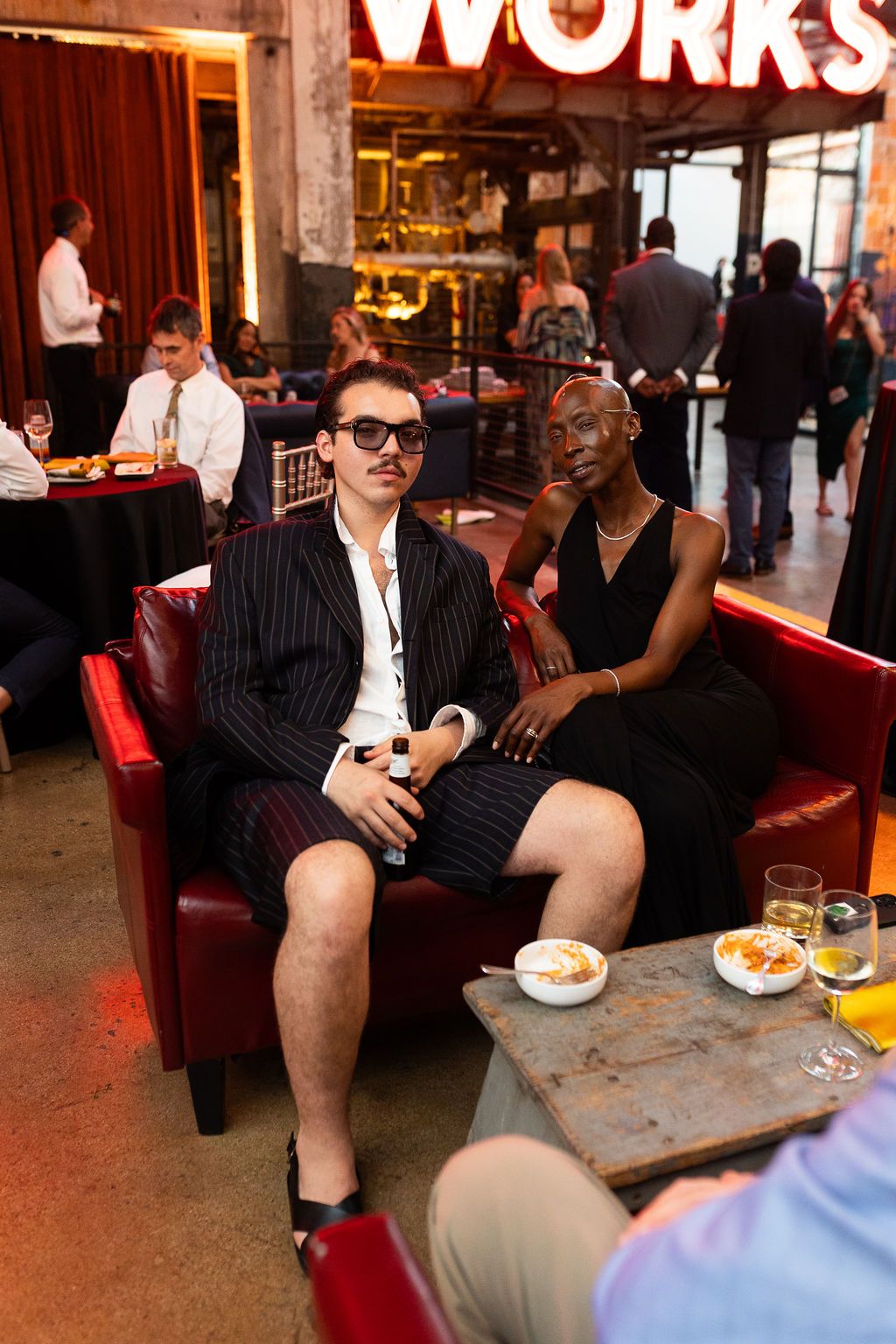Two wedding guests in stylish outfits sitting on red leather sofa at a reception venue.