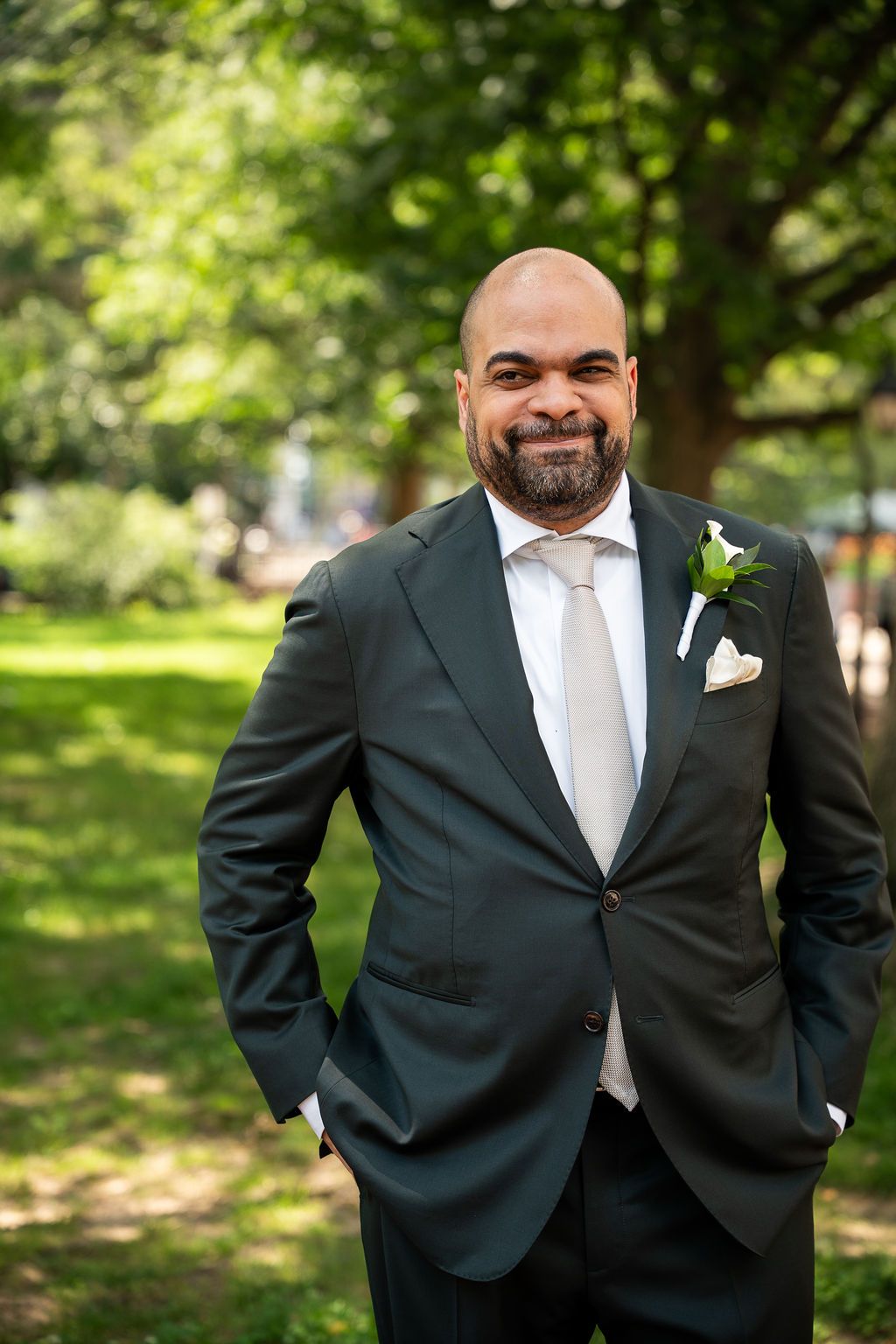 Groom in dark suit with white boutonniere smiling with hands in pockets in sunlit outdoor setting.