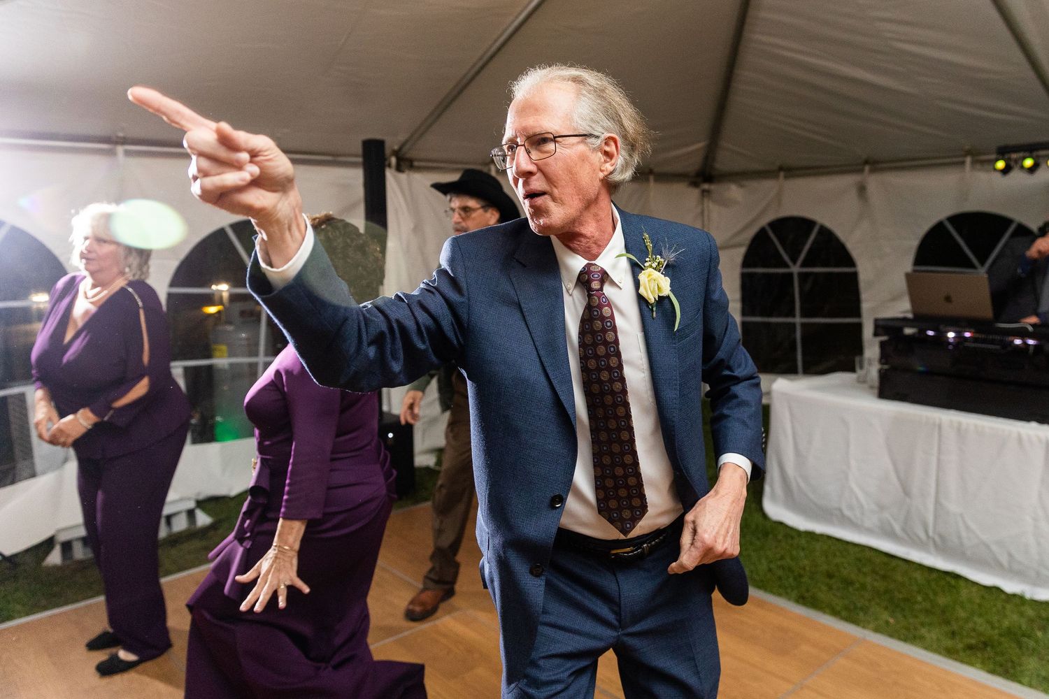 Lively older man in blue suit dances energetically at outdoor tented wedding reception celebration.