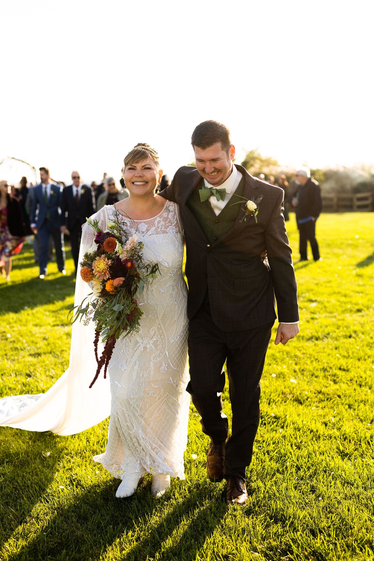 Smiling bride in lace gown holds autumn bouquet while walking with groom in green bow tie at fall wedding.