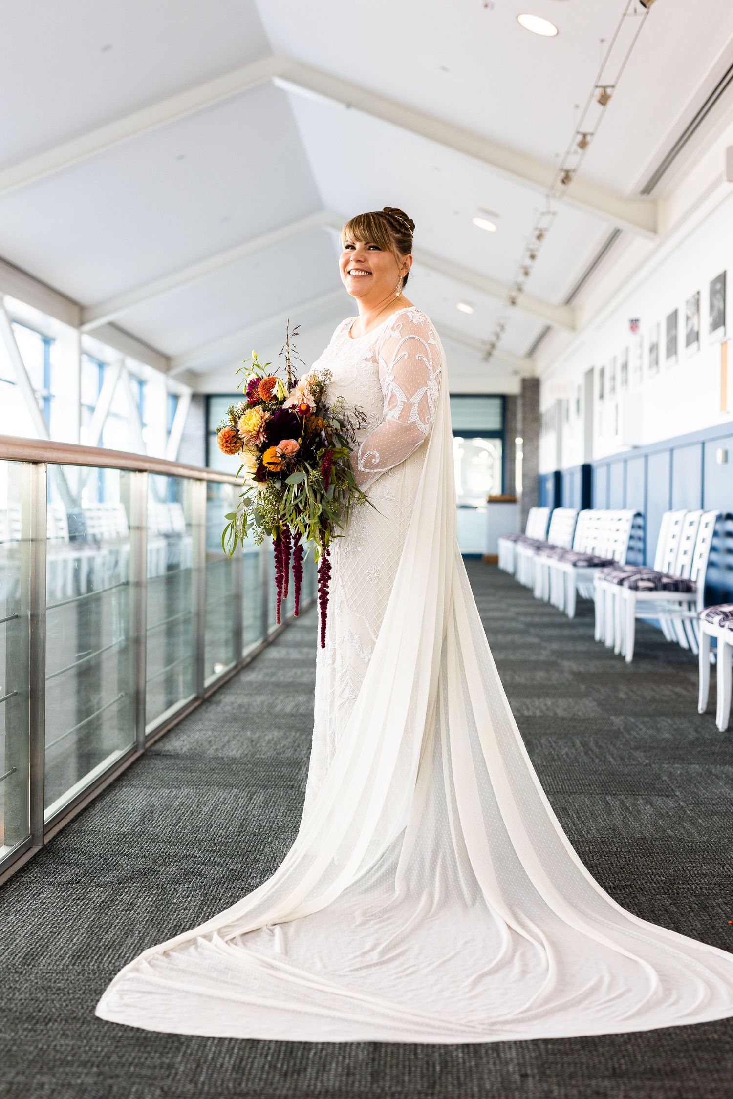Stunning bride in white lace gown with dramatic train poses in sunlit venue hallway with bouquet.