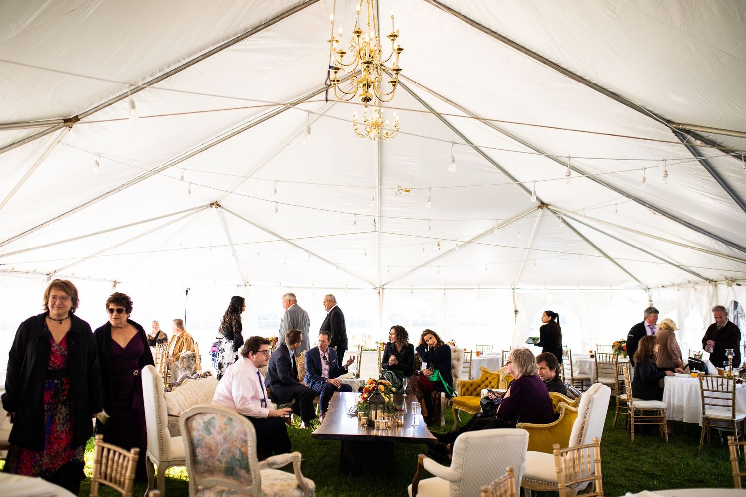 Elegant outdoor wedding reception under a white tent with chandeliers, guests mingling around decorated tables.
