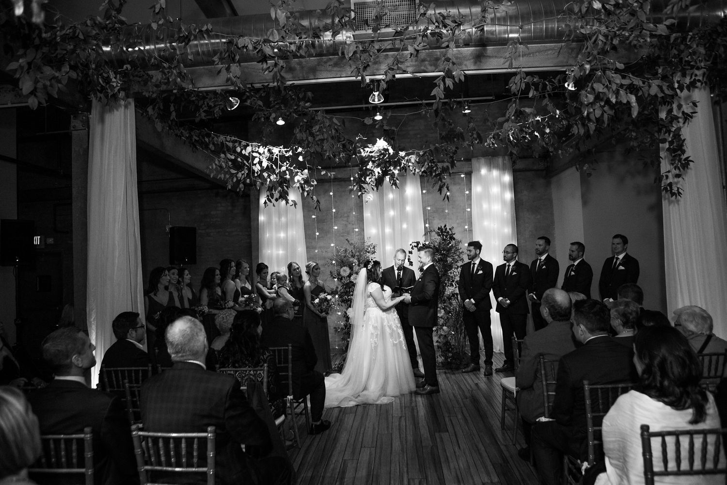 Black and white photo of romantic indoor wedding ceremony with greenery canopy overhead.