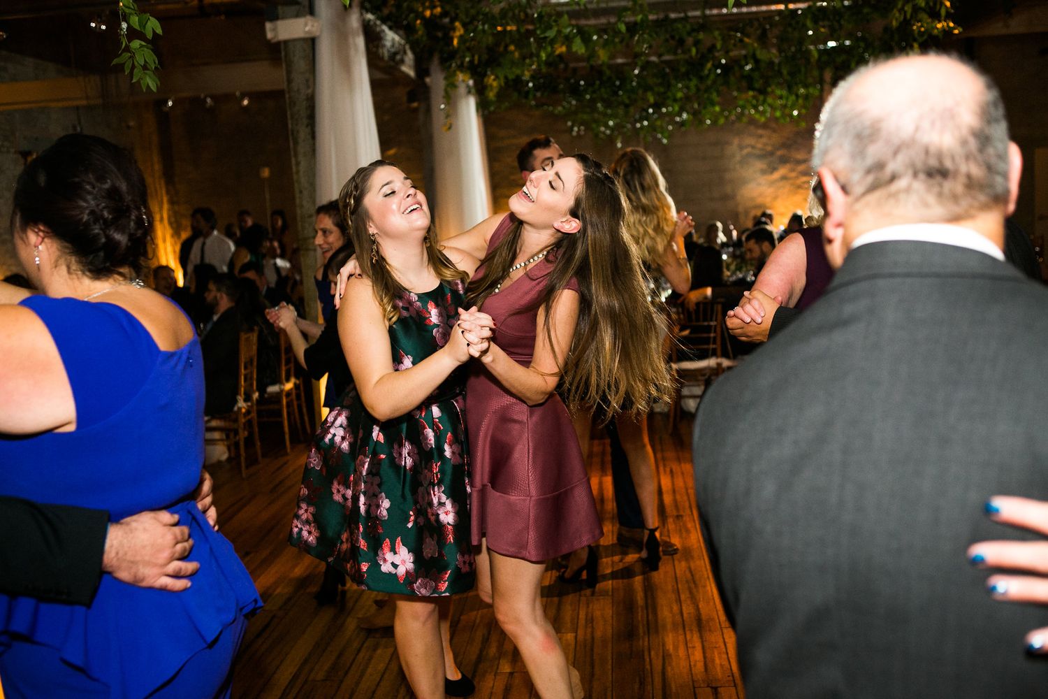 Two women in floral and burgundy dresses dancing and laughing at wedding reception.