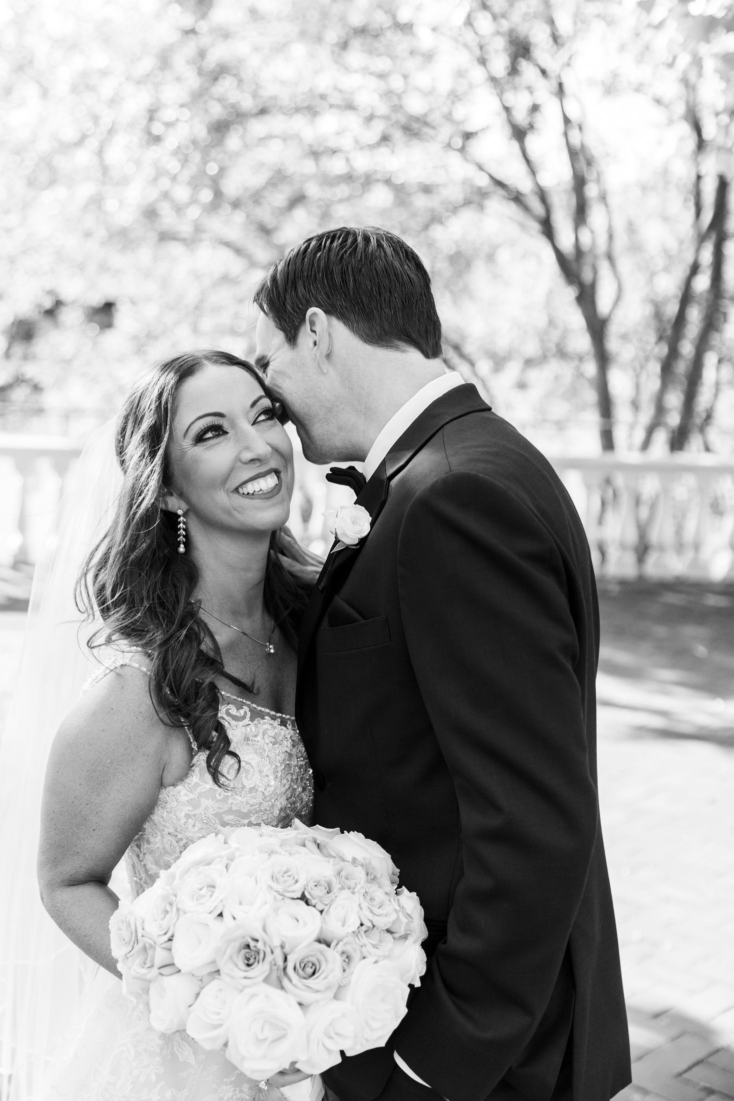 Black and white photo of a smiling bride holding roses while groom kisses her cheek outdoors.