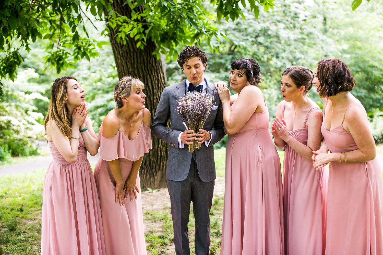Groom in gray suit holds lavender bouquet while four bridesmaids in pink dresses react with delight.