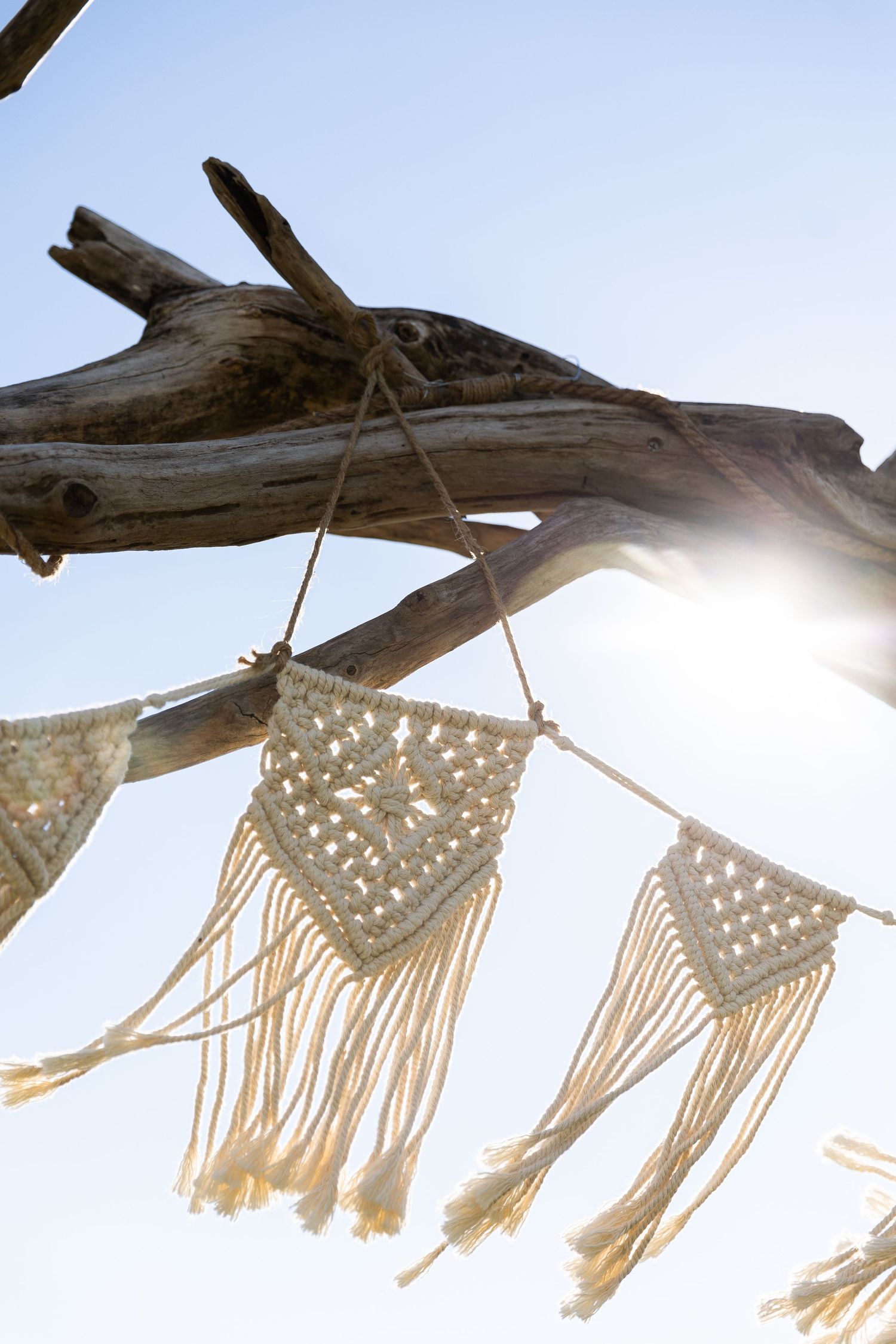 Boho macrame bunting hanging from driftwood branch with sun flare against blue sky.