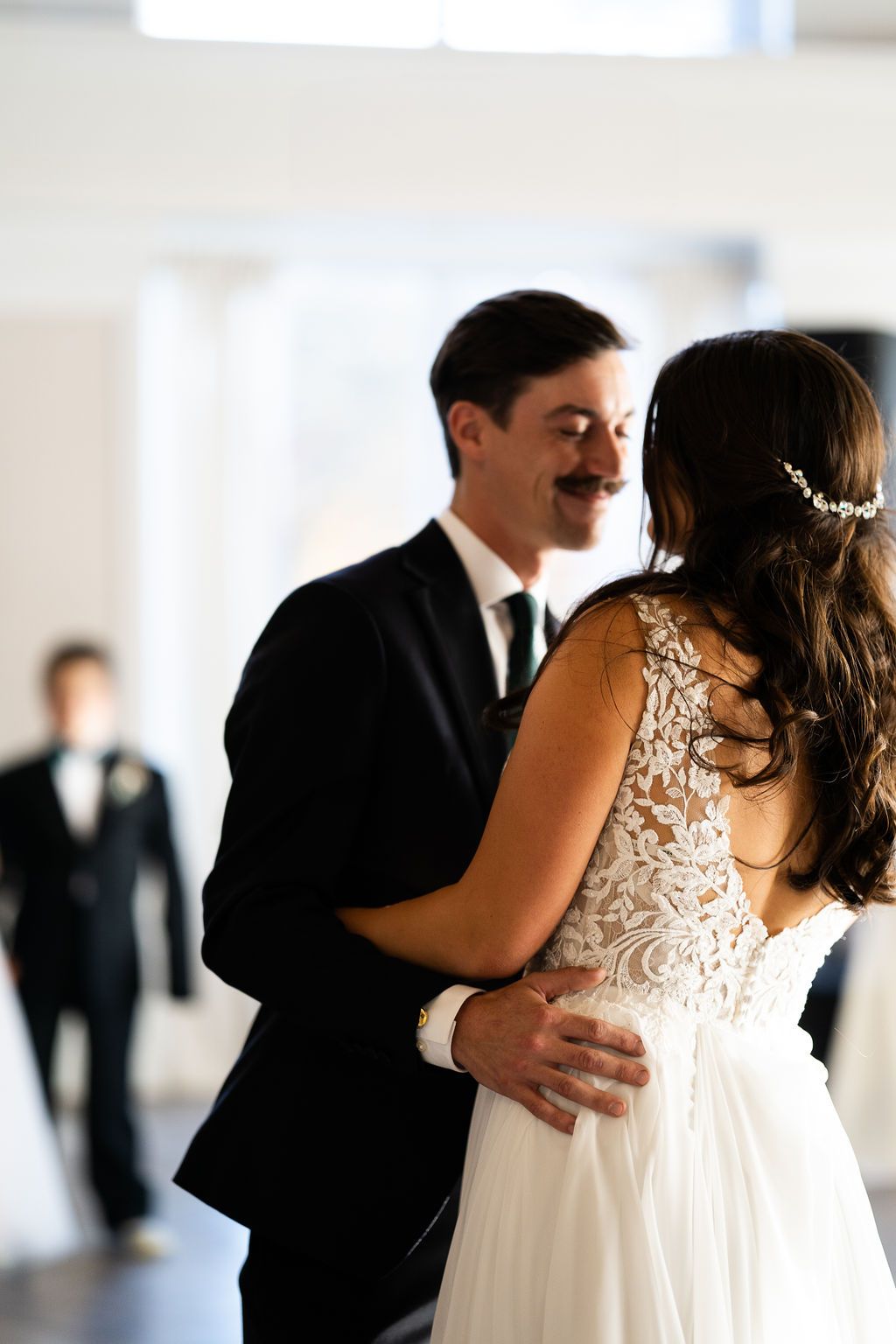 Groom smiles lovingly at bride during first dance, showcasing intricate lace back detail of her wedding gown.