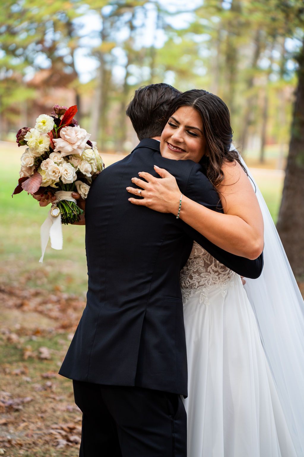 Smiling bride in lace wedding gown embraces groom holding a bouquet of white and red flowers.