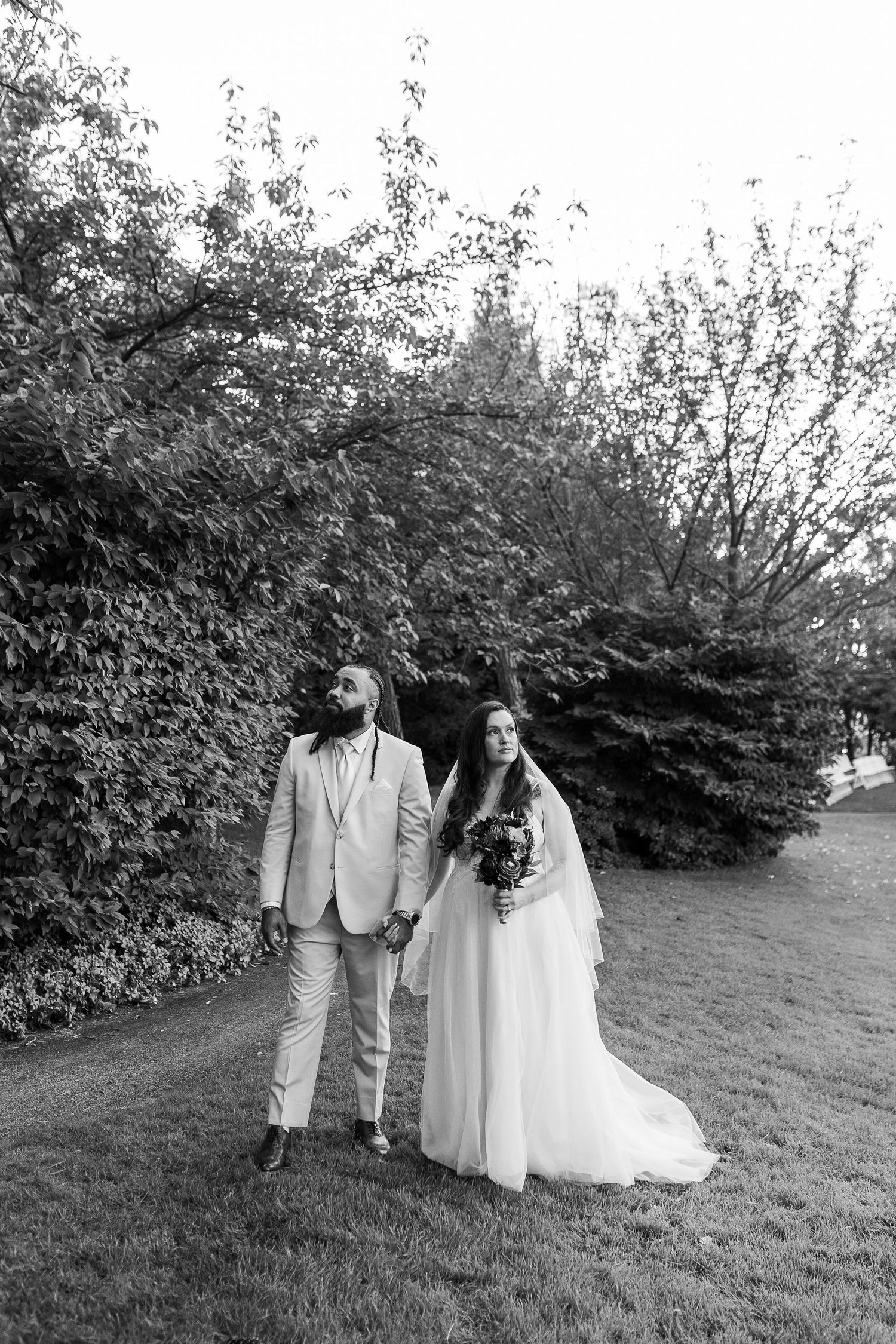 Black and white photo of bride and groom walking hand in hand through a lush garden path on their wedding day.