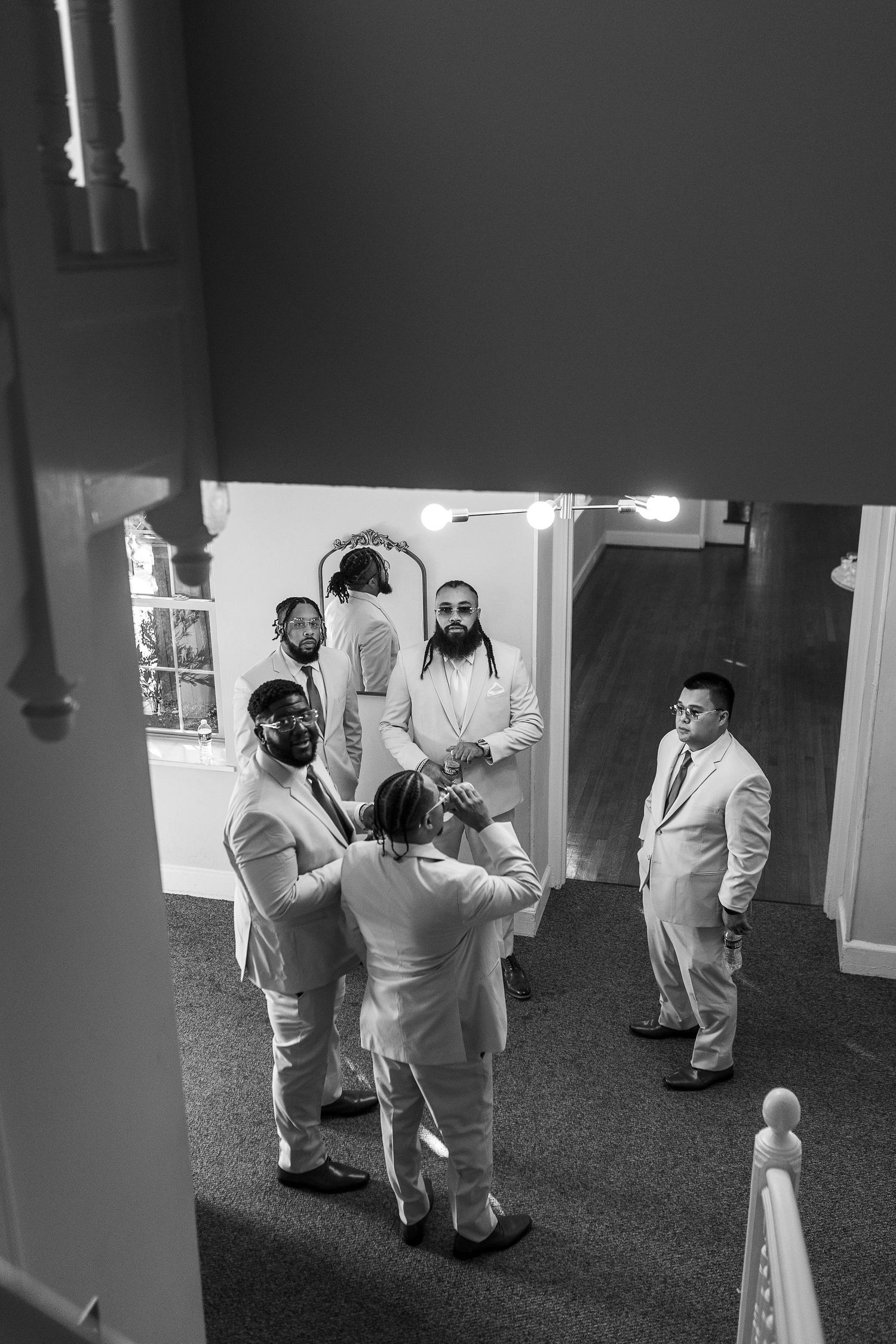 Black and white overhead view of groomsmen in white suits helping each other get ready indoors.