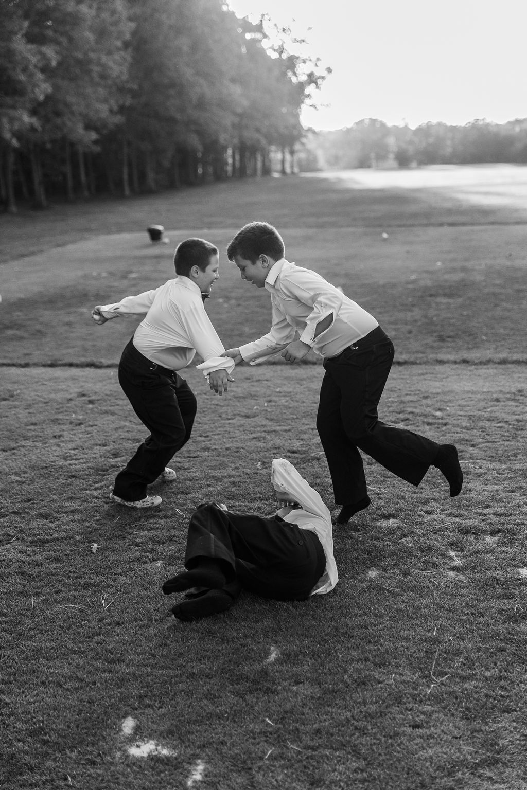 Black and white photo of two groomsmen playfully wrestling on golf course lawn at wedding.