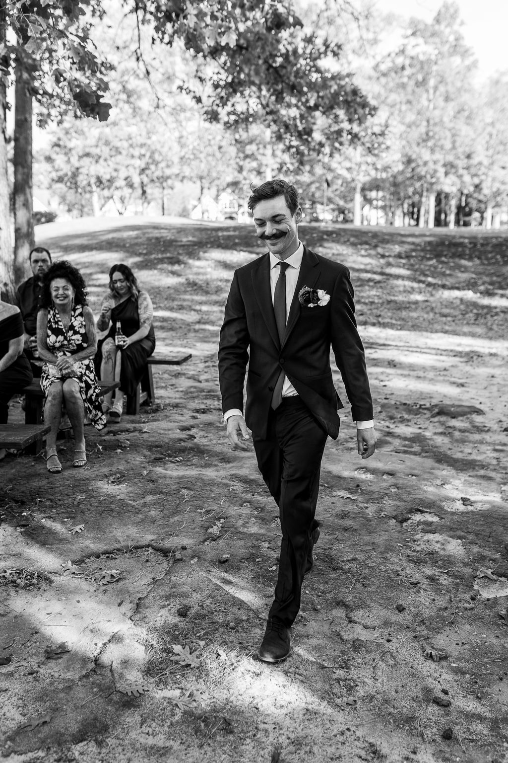 Groom in black suit with boutonniere walks down aisle at outdoor wedding ceremony, guests seated in background.