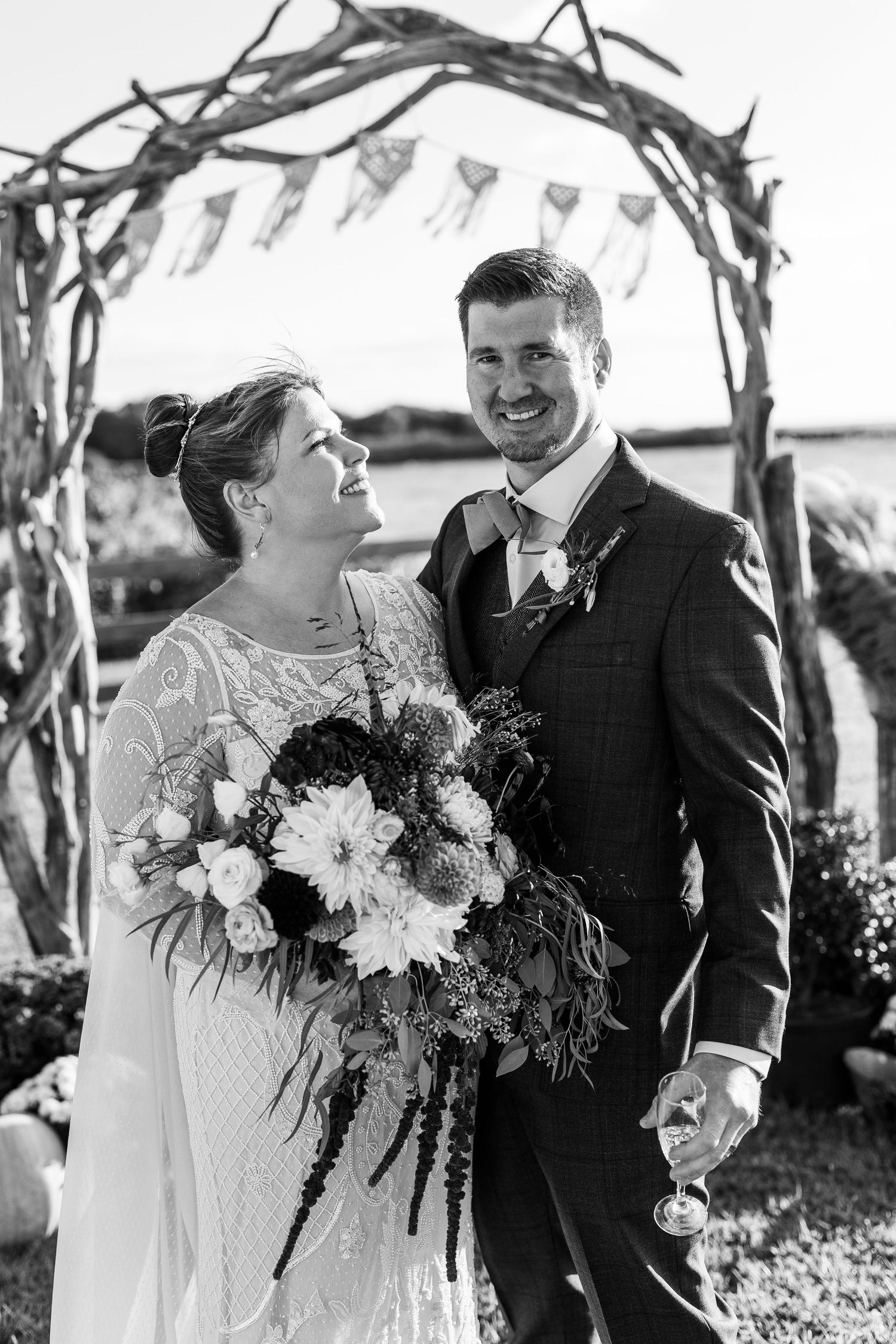 Black and white portrait of smiling couple posing under rustic driftwood wedding arch with lush floral bouquet.