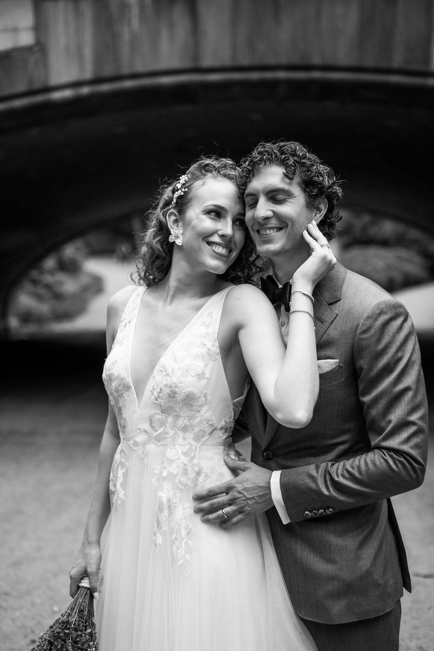 Black and white close-up of smiling bride and groom embracing beneath a stone bridge.