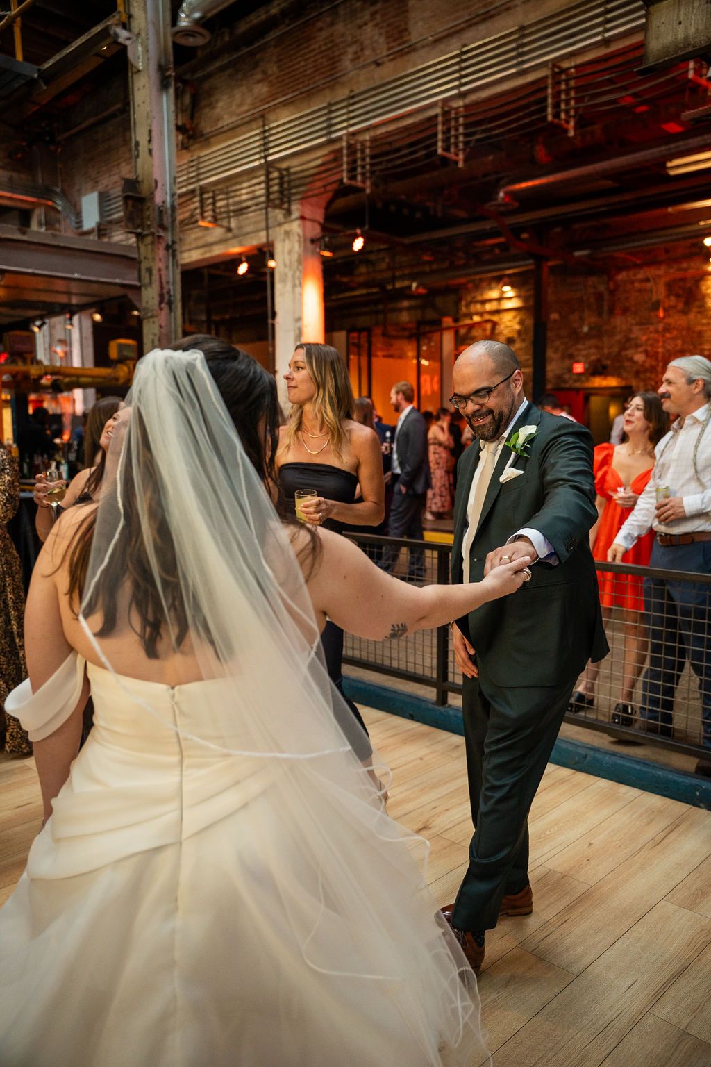 Groom spins bride in wedding dress on dance floor surrounded by guests at rustic industrial venue.