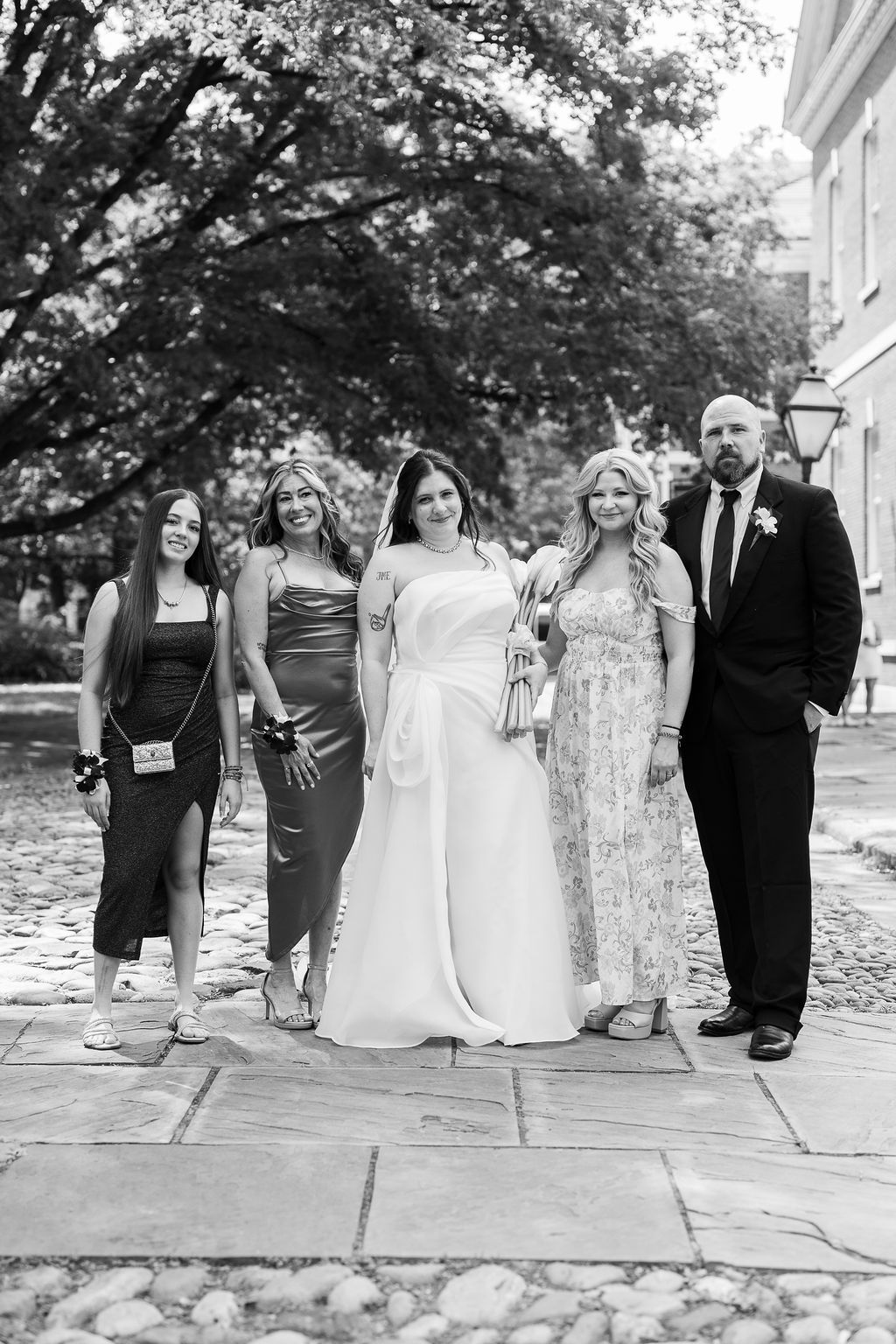 Bride poses with four guests in elegant attire on cobblestone path in black and white photo.