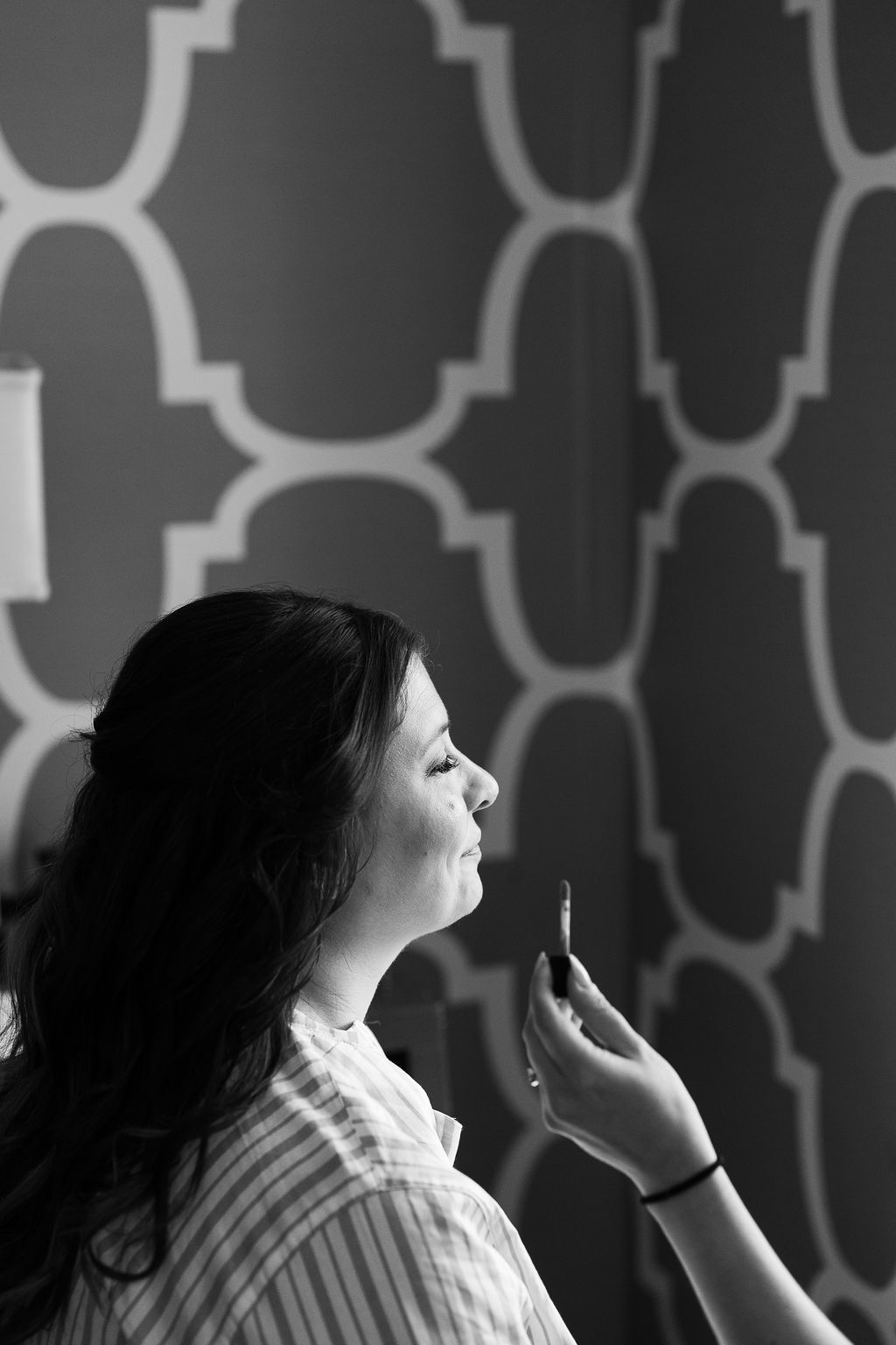 Black and white profile portrait of a bride smiling during makeup application against patterned wallpaper.