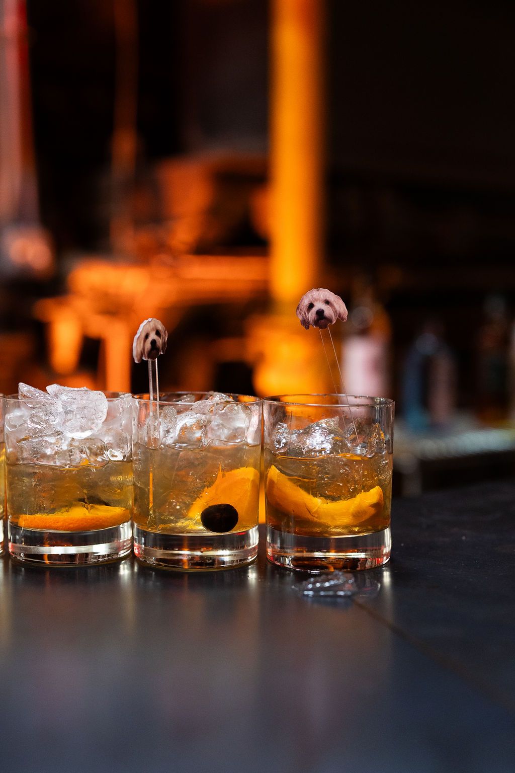 Three old fashioned cocktails with orange slices and cherry garnishes on a bar.