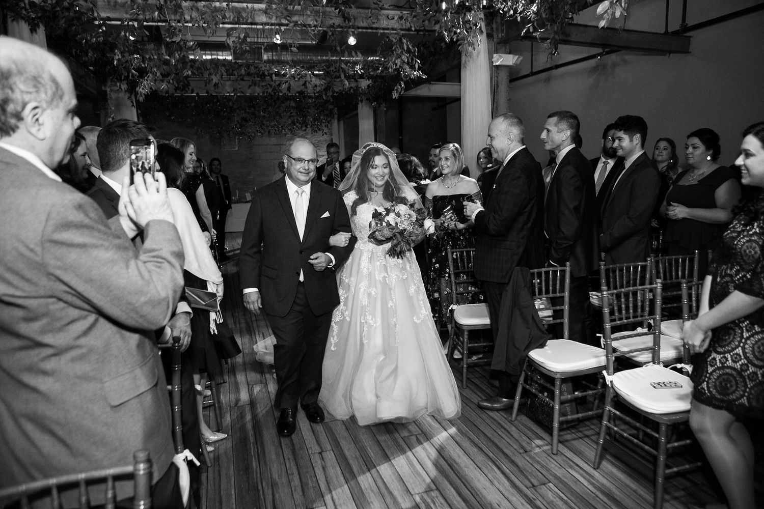 Black and white photo of bride walking down aisle with escort at indoor wedding ceremony.