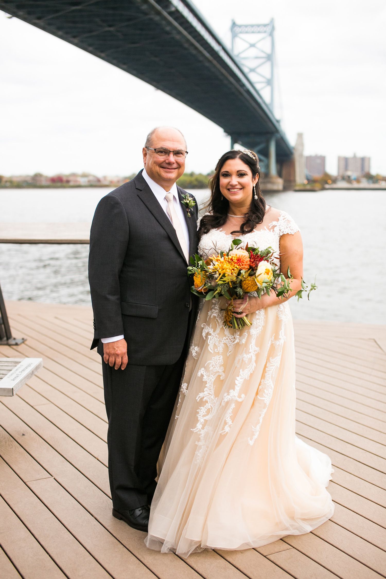 Bride and groom pose on waterfront deck with Benjamin Franklin Bridge backdrop on wedding day.