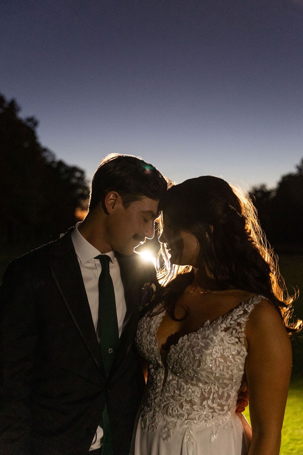Bride and groom sharing romantic moment at twilight with glowing backlight between them.