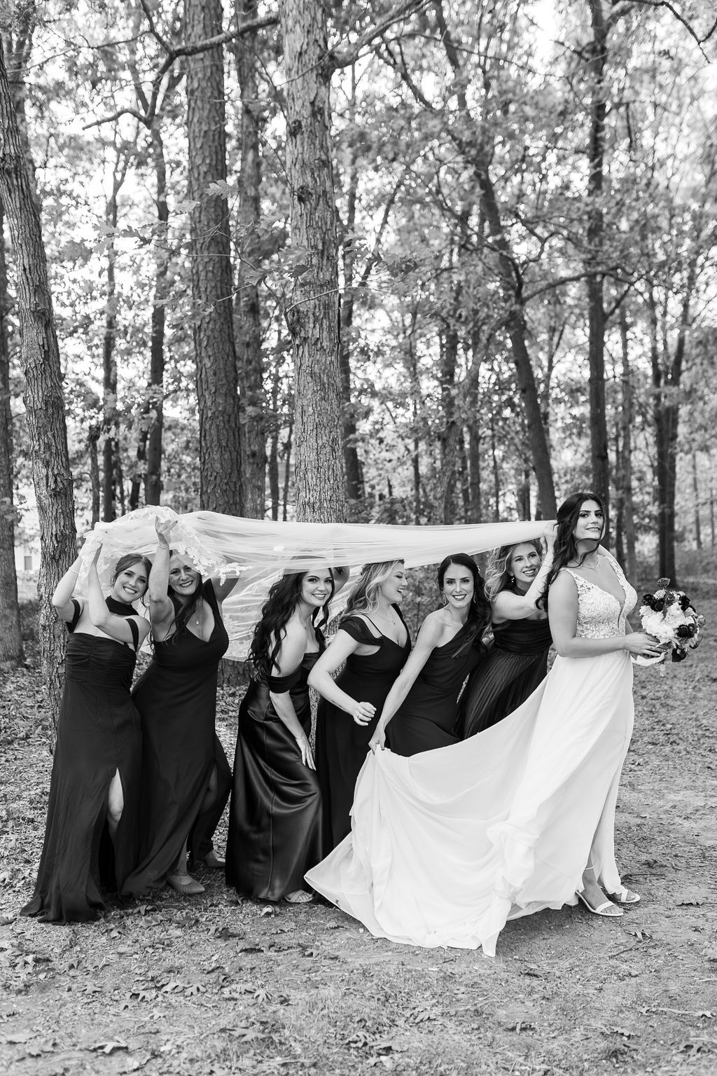 Bridesmaids in dark gowns huddle under bride's veil in a wooded setting, captured in black and white.