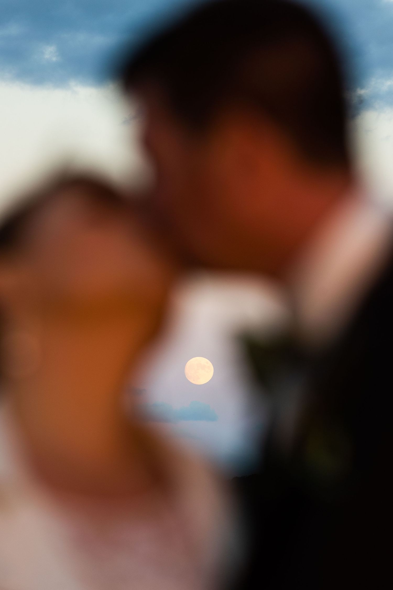Soft focus color photo of bride and groom kissing with full moon glowing between them.