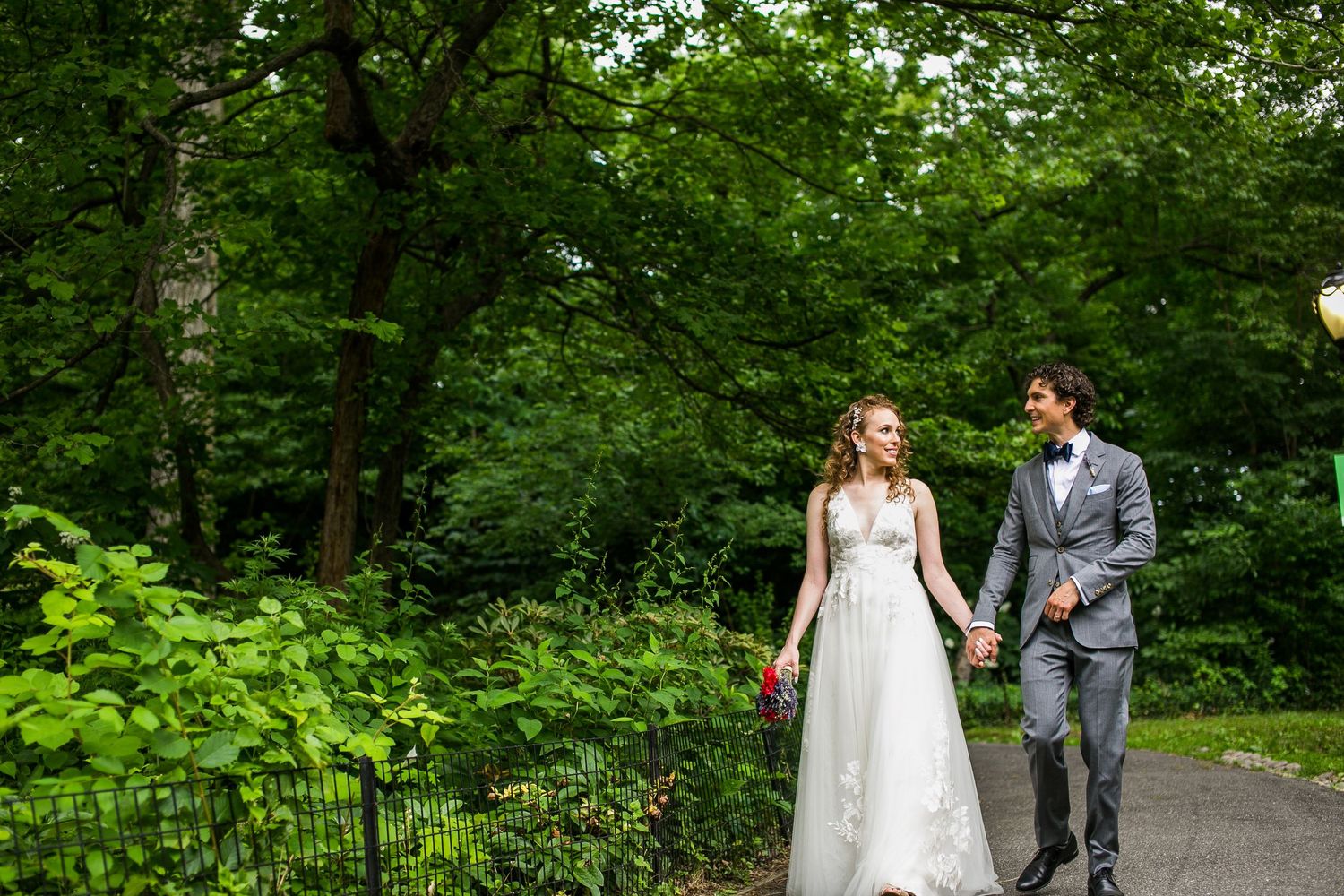 Bride and groom walk hand in hand through lush green Central Park on their wedding day.