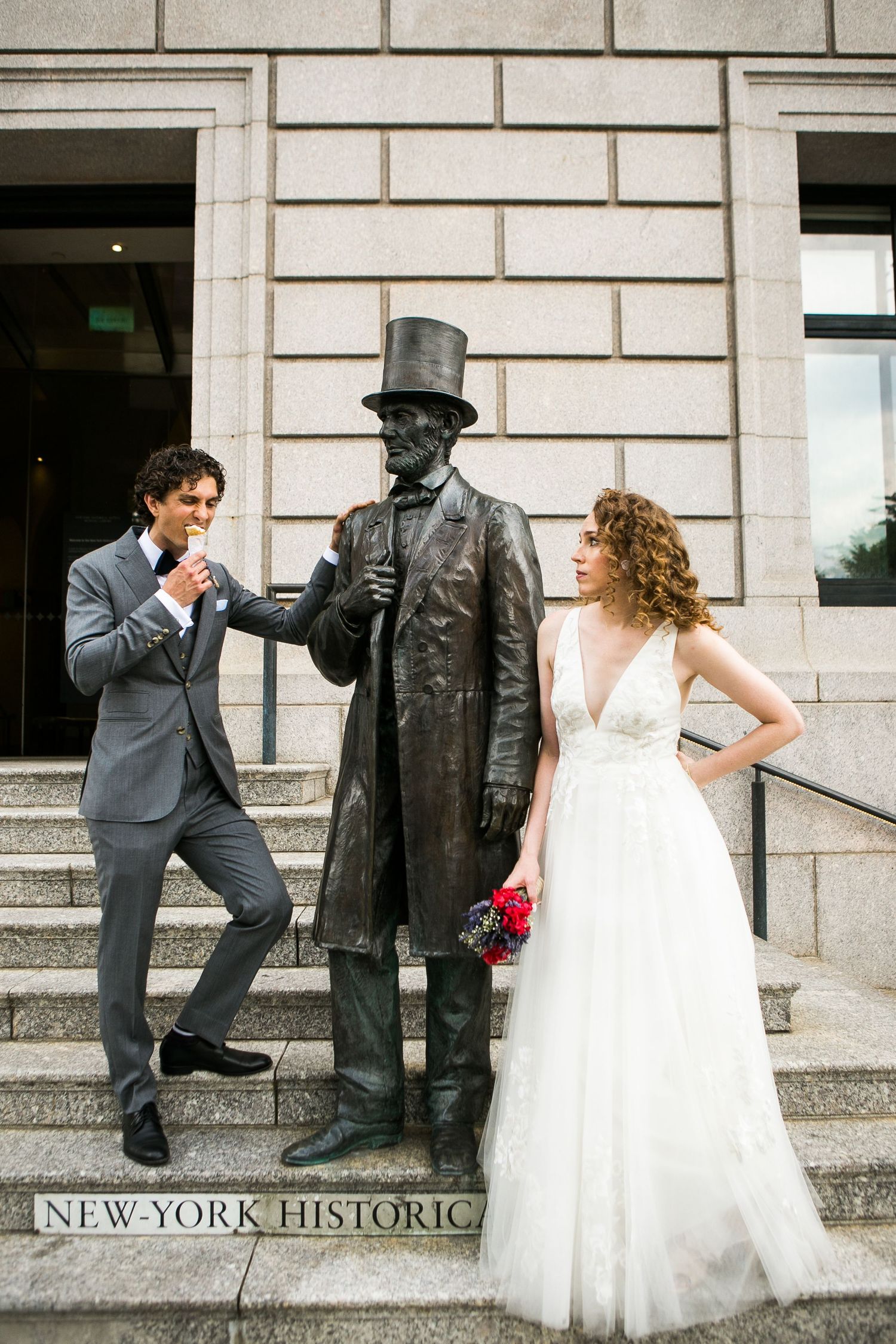 Bride and groom posing with a bronze statue on the steps of the New-York Historical Society building.