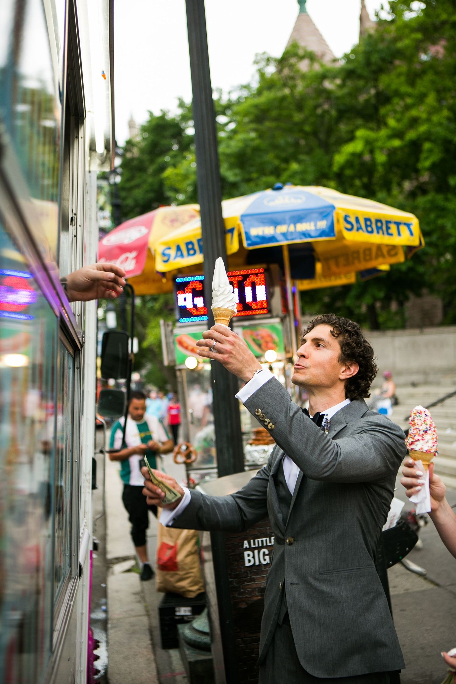 Groom in a gray suit receiving soft-serve ice cream cones from a street vendor truck near Sabrett stands.