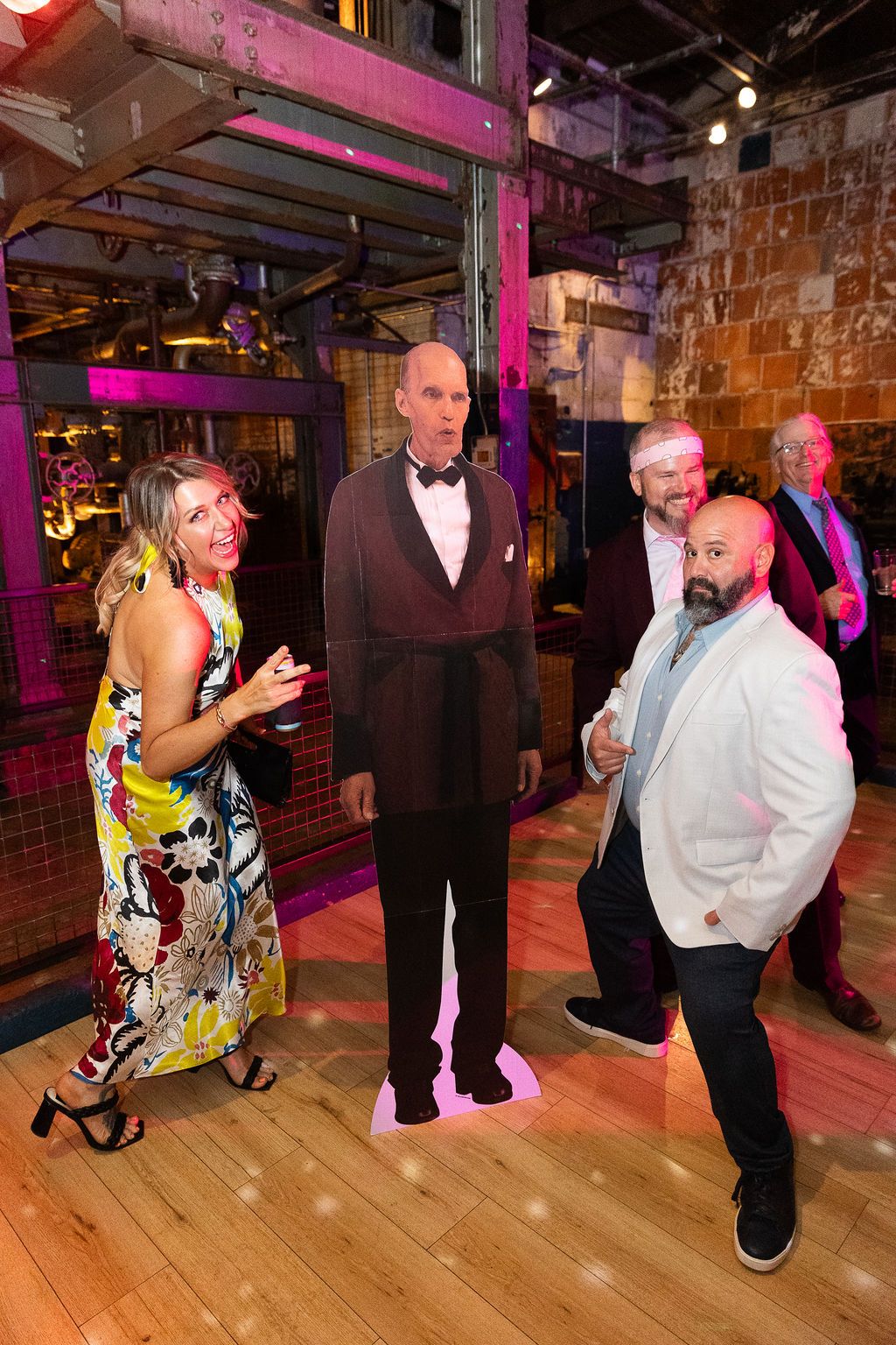 Two guests interact with a life-size cardboard cutout of a man in formal attire at an indoor event venue.