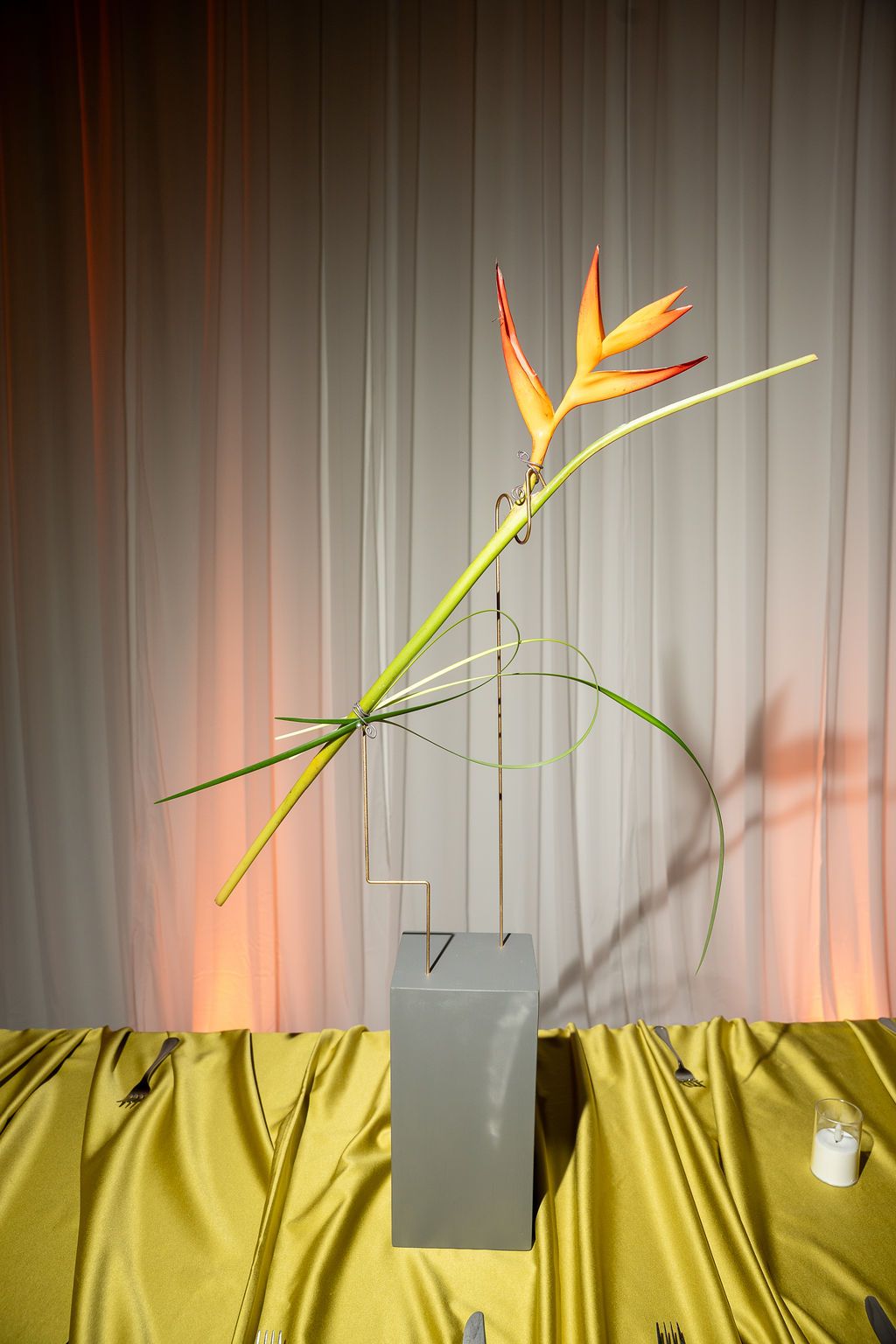 Elegant bird of paradise flower arrangement in a gray vase on a gold satin tablecloth against a draped backdrop.