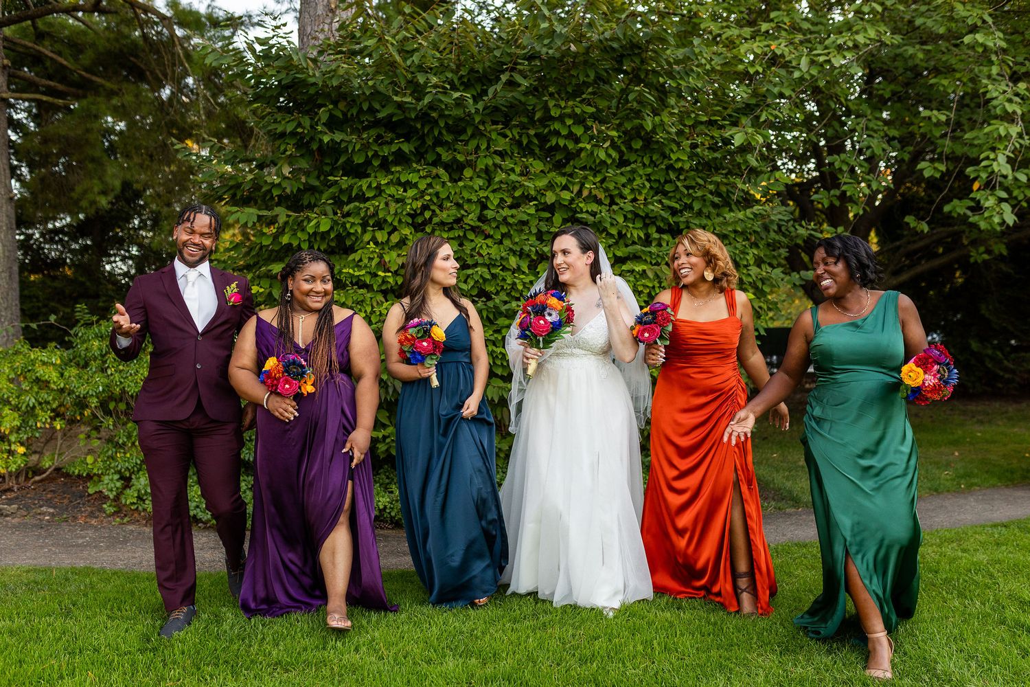 A bride in white walks with five bridesmaids in colorful mismatched gowns on a green lawn.