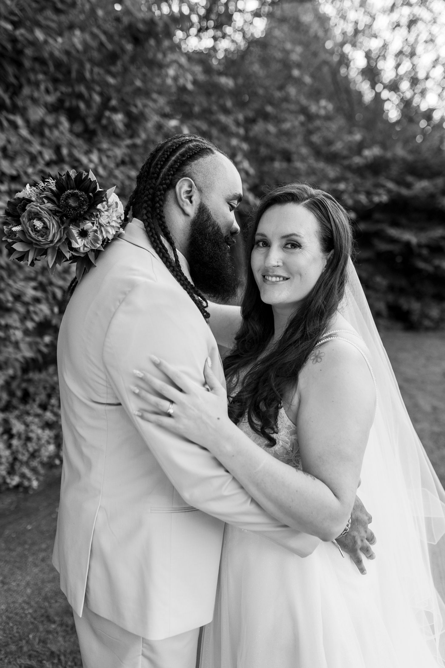 Black and white portrait of smiling bride embracing bearded groom in a lush garden setting.