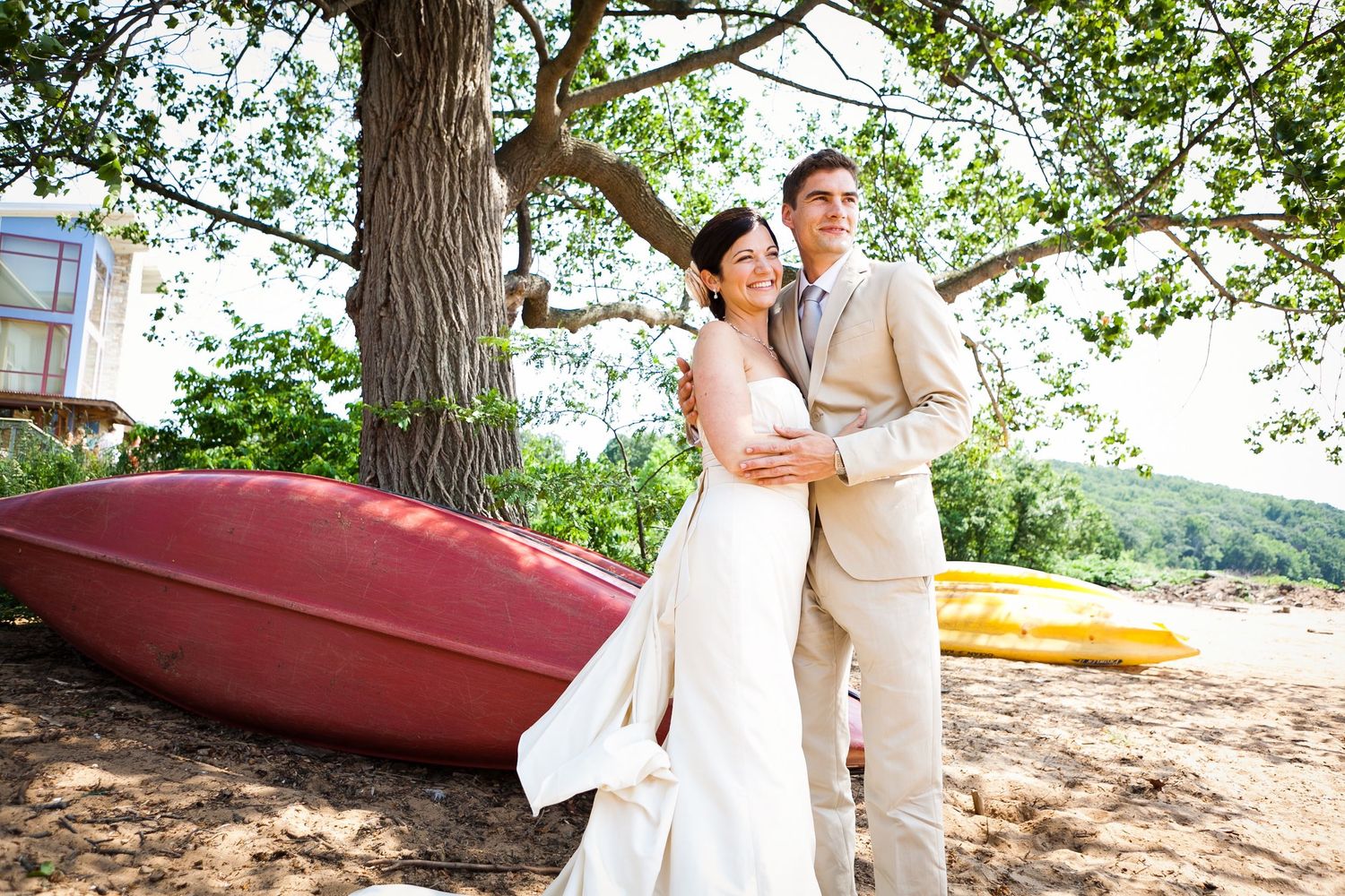 Bride and groom posing near colorful canoes under a tree outdoors.