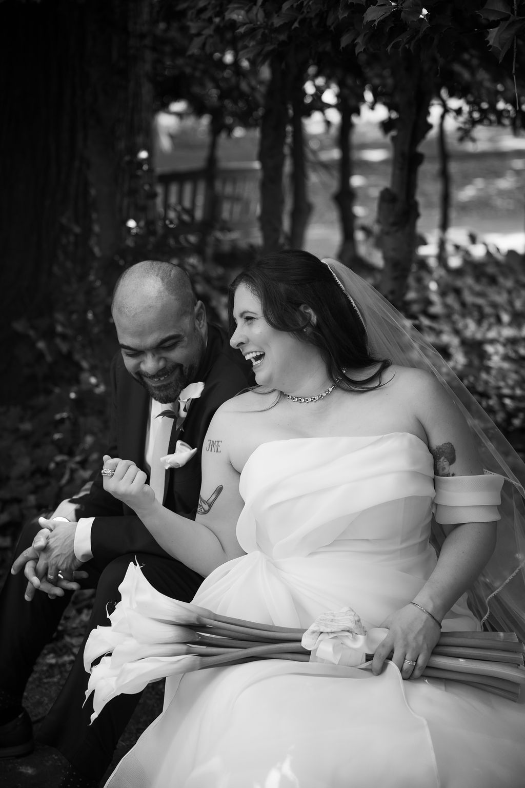 Black and white photo of laughing bride and groom sitting on bench, bride holding calla lily bouquet.