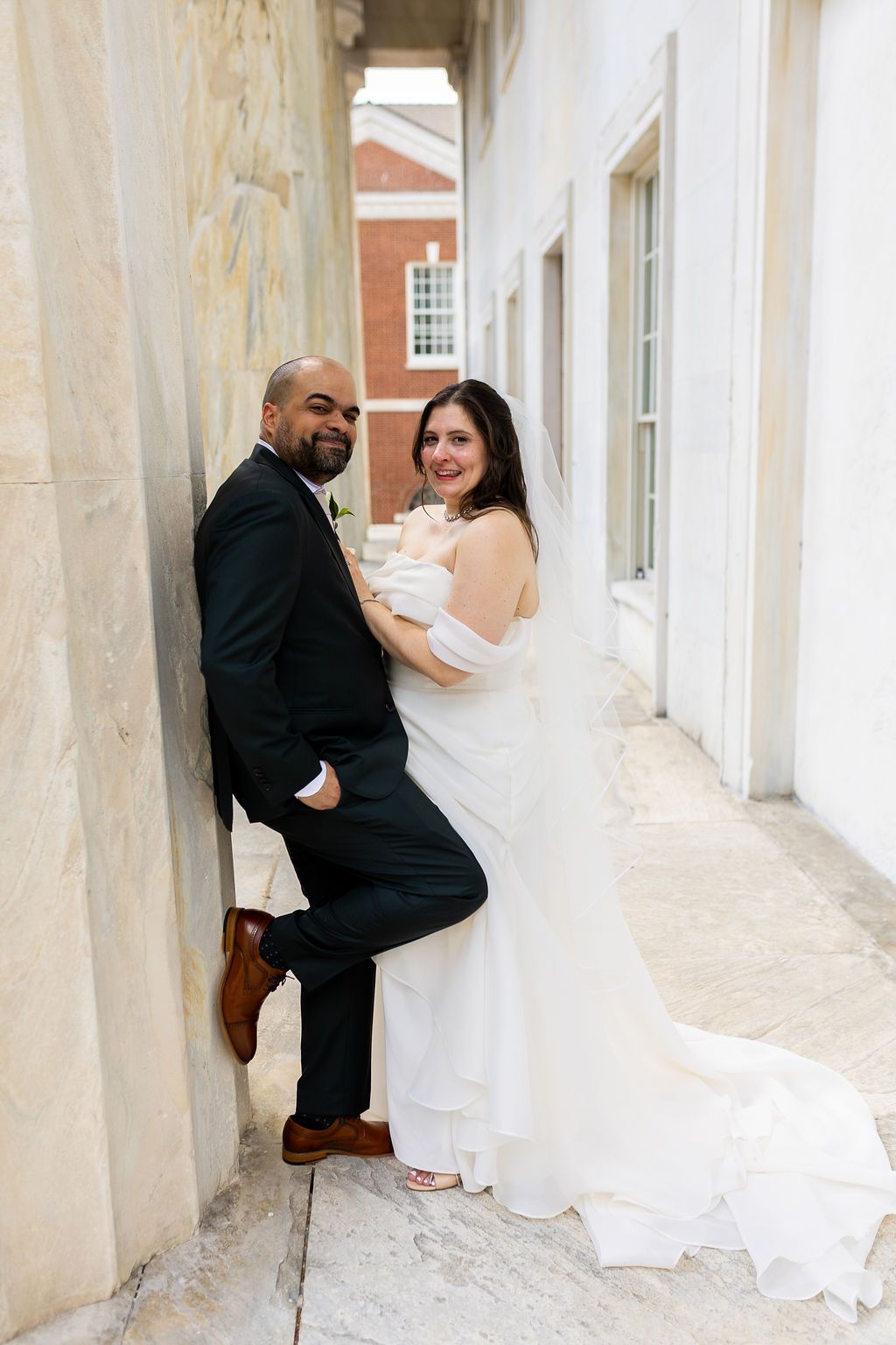 Smiling bride and groom posing together against a marble wall with a brick building backdrop.