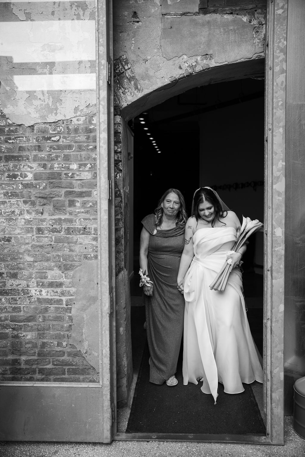 Black and white photo of bride in white gown and bridesmaid exiting brick doorway before ceremony.