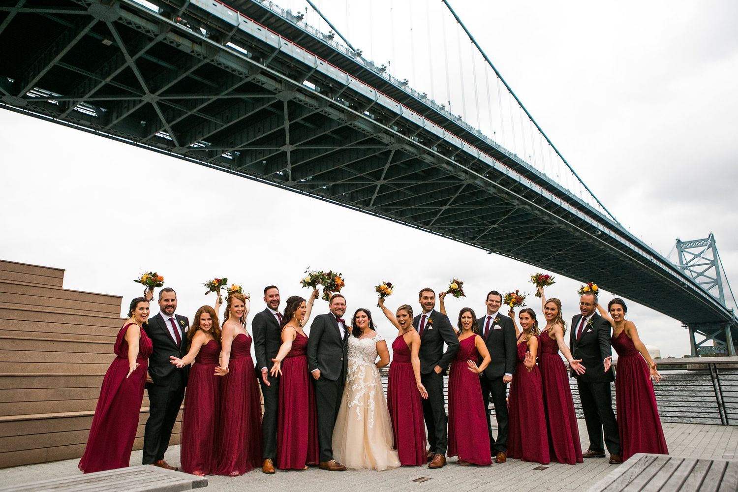 Large wedding party in burgundy dresses and black suits poses under the Benjamin Franklin Bridge on a sunny day.