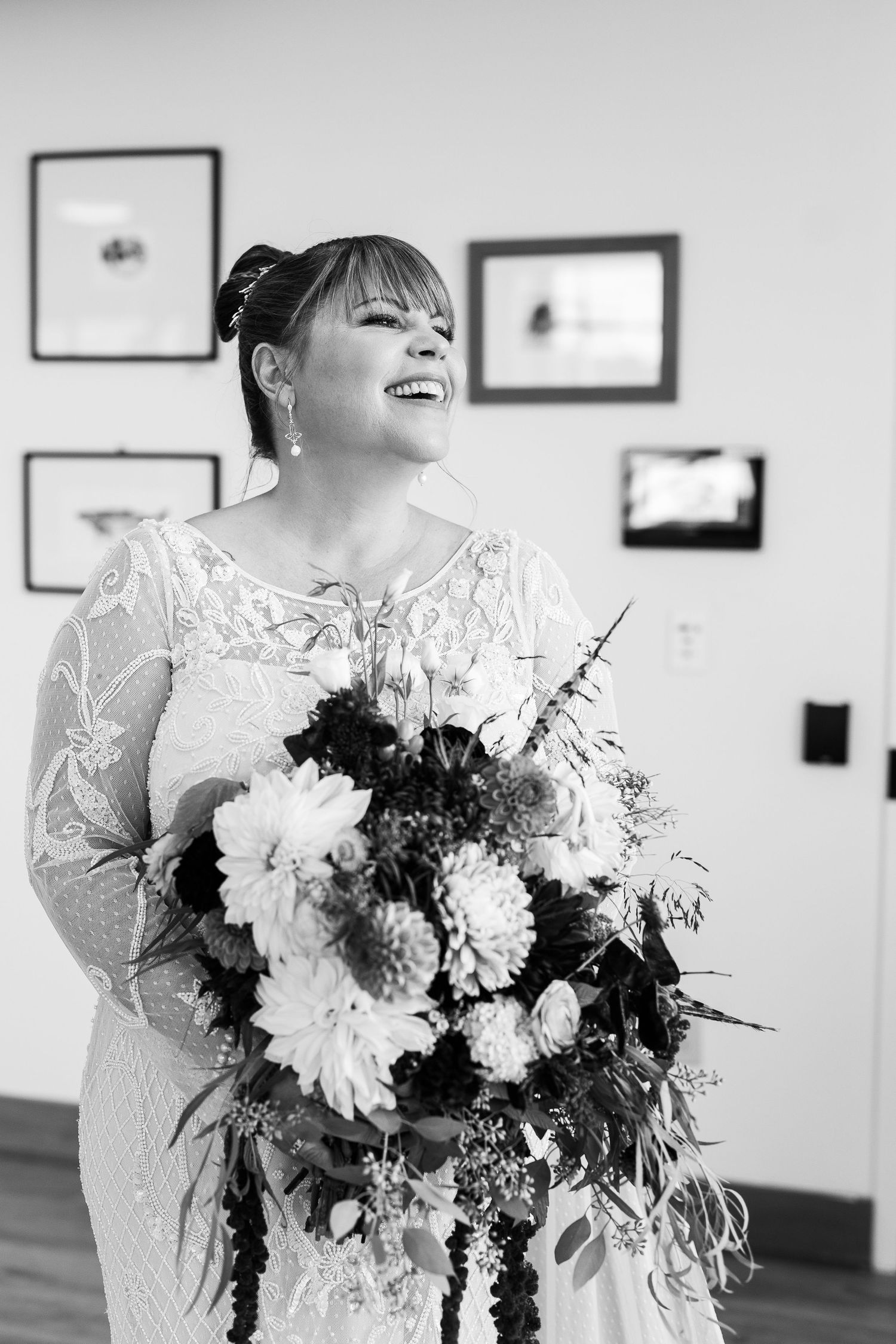 Black and white photo of smiling bride holding large floral bouquet in lace wedding gown indoors.