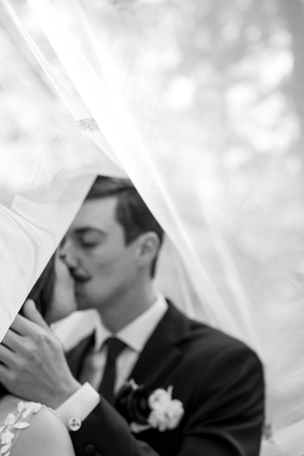 Black and white artistic photo of bride and groom kissing under wedding veil.