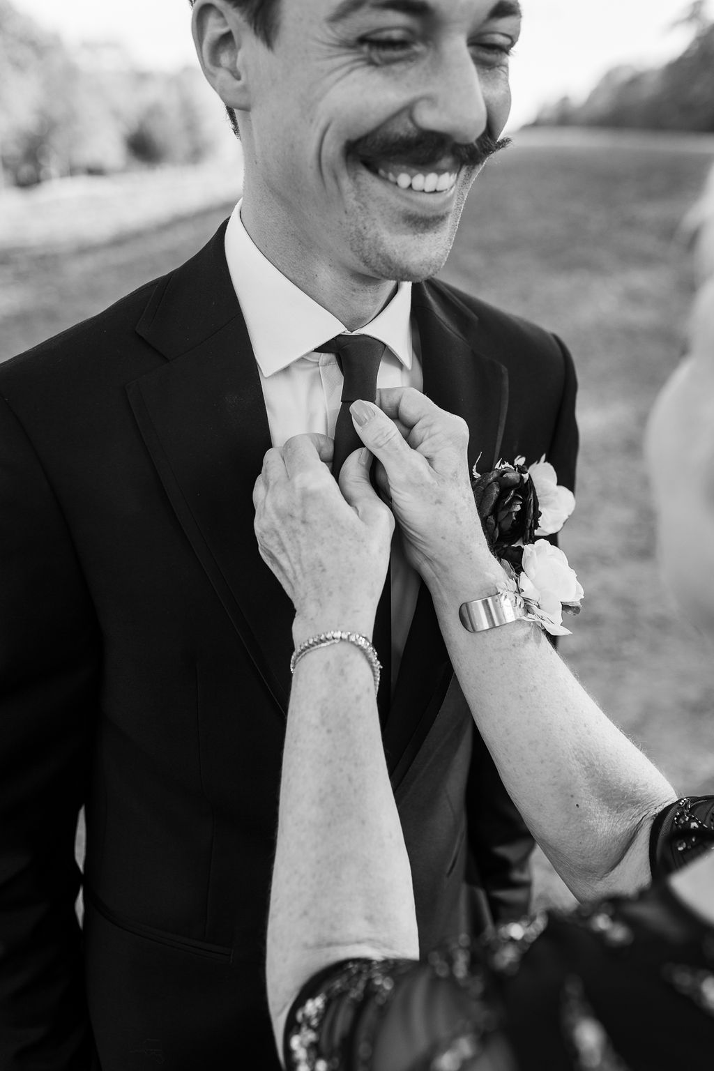 Black and white photo of groom with mustache smiling as hands adjust his tie at wedding.