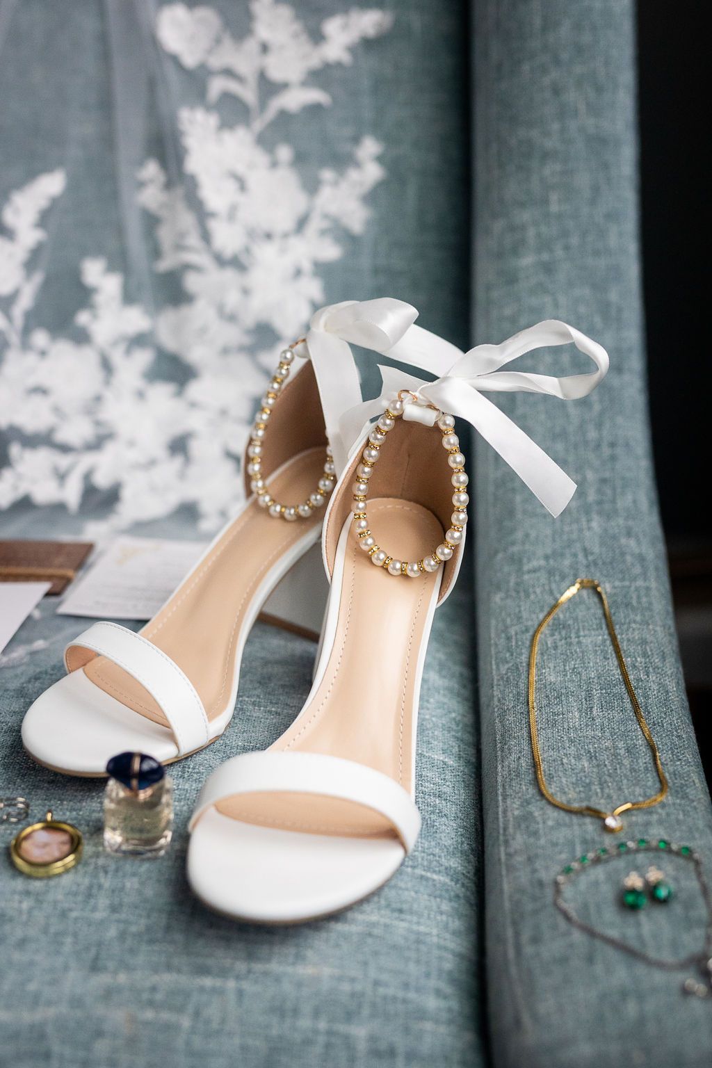 White bridal heeled sandals with pearl ankle straps and ribbon ties displayed on a blue linen surface.