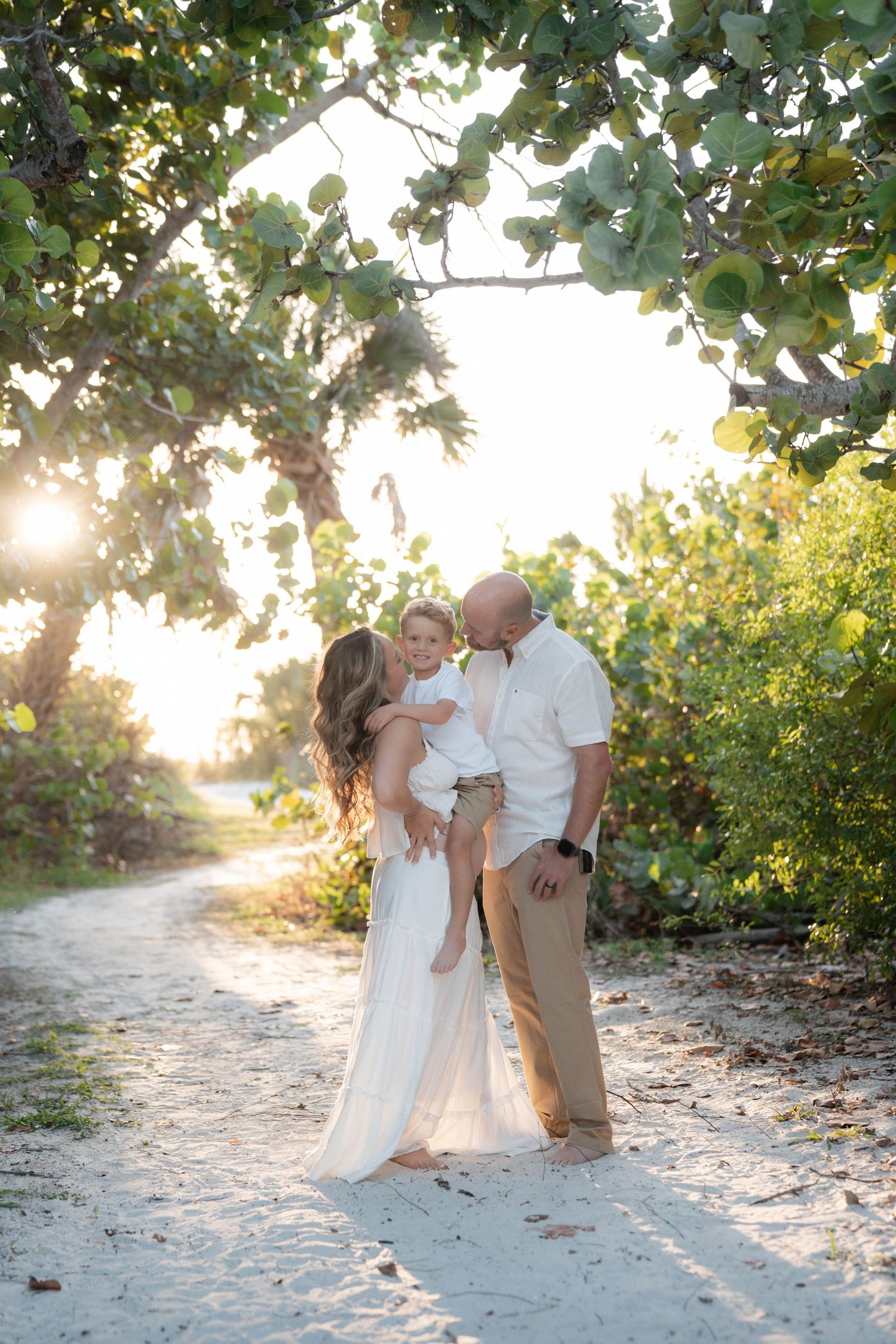 Family of three sharing a tender moment on a sandy beach path at golden hour, surrounded by lush tropical greenery.