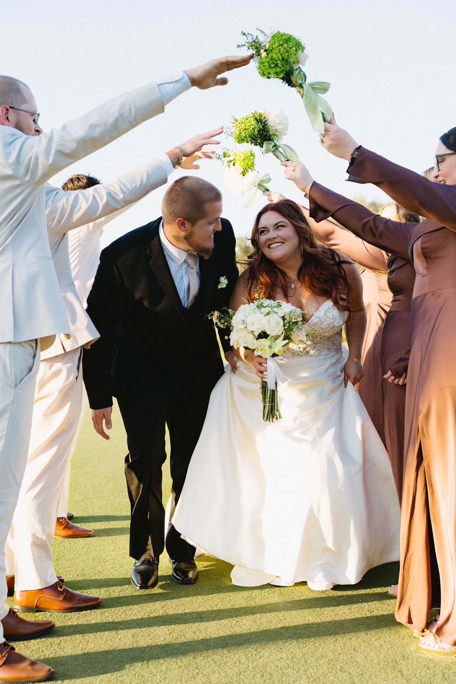 Bride and groom walking through a bouquet arch formed by their smiling wedding party.