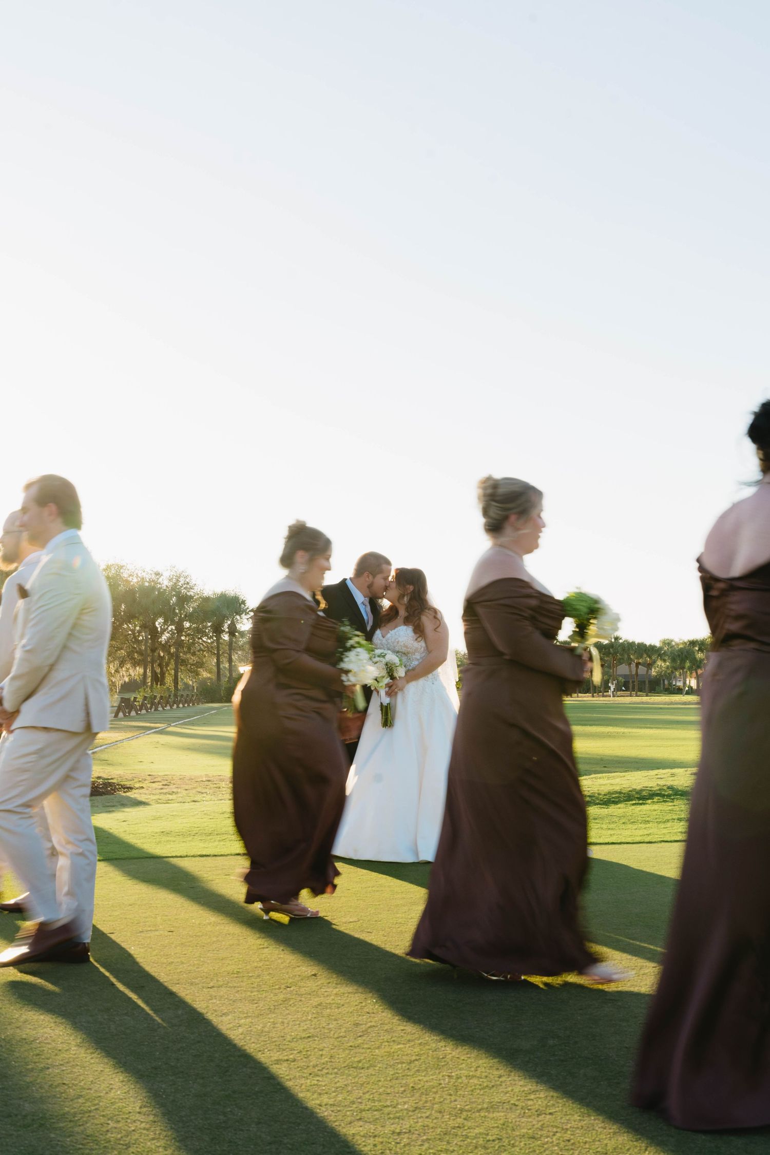 Wedding party gathered on a green golf course at golden hour with bridesmaids in dark gowns and couple kissing.