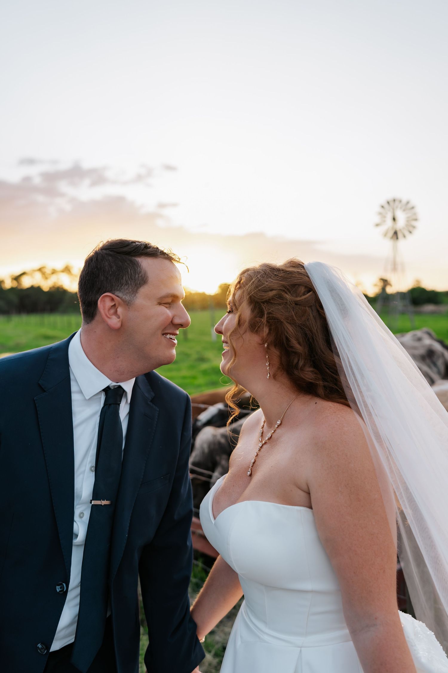 Bride and groom smile at each other during golden hour with windmill backdrop on farm.