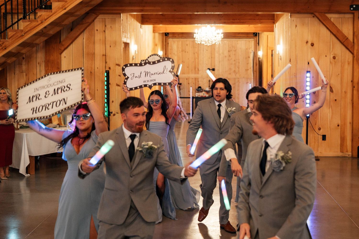 Wedding party celebrates grand entrance with glowing foam sticks in rustic barn venue.
